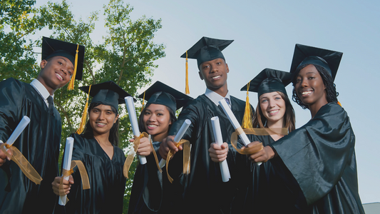Group of diverse graduates wearing black caps and gowns with gold tassels, smiling and holding rolled diplomas tied with golden ribbons, standing outdoors with trees and a clear blue sky in the background.