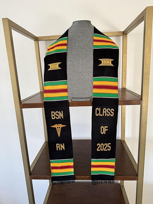 A black and yellow BSN RN Kente graduation stole displayed on a grey mannequin torso, focusing on the chest and shoulder area to showcase how it would look when worn. The stole is placed on top of a wooden shelf. The stole features the bold vertical 'BSN RN' lettering on the left, with a medical logo between 'BSN' and 'RN', and 'CLASS OF 2025' embroidered horizontally on the right. The custom Kente pattern includes green, yellow, red, and green stripes in three sets on each side, with fabric made of rayon."