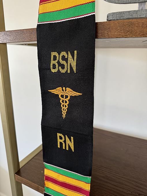 A close-up image of a black and yellow BSN RN Kente graduation stole, zoomed in on the bold vertical 'BSN RN' lettering on the left, with a medical logo placed between 'BSN' and 'RN'. The stole is displayed on a grey mannequin torso, positioned on top of a wooden shelf.