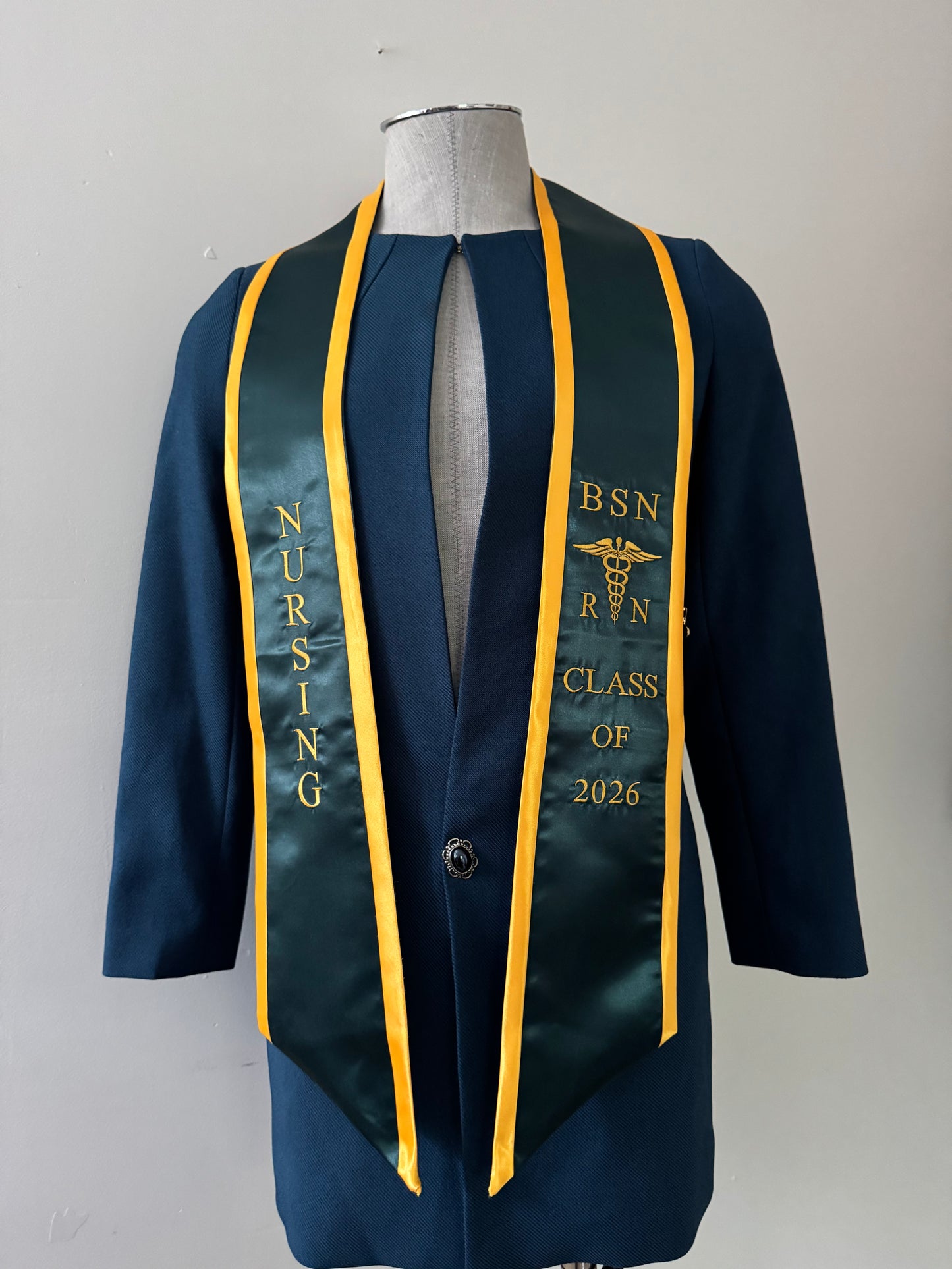 A forest green with gold trim BSN RN graduation stole, displaying bold 'BSN RN' lettering embroidered on the left, a medical logo with BSN RN on the right, and 'CLASS OF 2026' embroidered horizontally below. The stole has slanted tips and a white plain background. All text is embroidered.