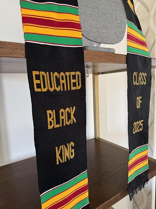 A black graduation stole with Kente cloth patterns in red, yellow, and green, draped over a gray mannequin. The stole features gold embroidery with the words "EDUCATED BLACK KING" on one side and "CLASS OF 2025" on the other, along with gold pillar symbols.