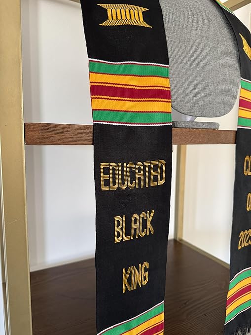 A beautifully woven black graduation stole with colorful Kente patterns, featuring gold embroidery that reads "EDUCATED BLACK KING" and "CLASS OF 2025." The stole is displayed on a fabric-covered mannequin against a neutral background.