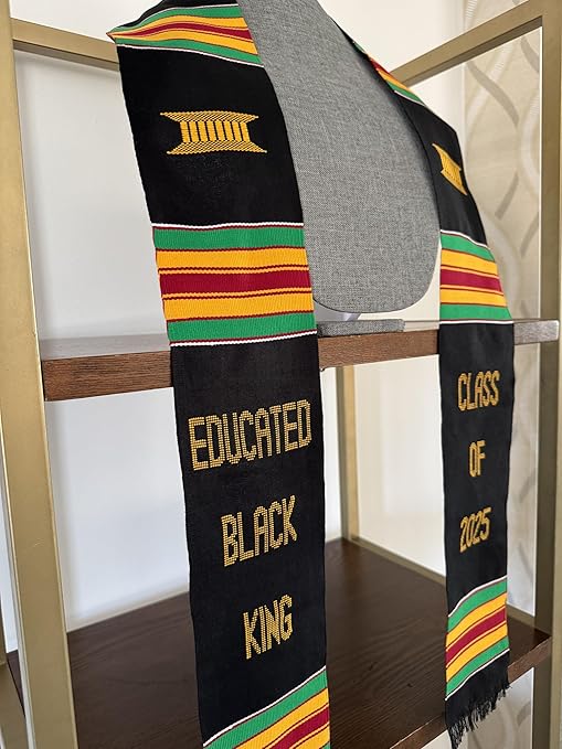 A black graduation stole with Kente-inspired stripes, featuring gold embroidered text that reads "EDUCATED BLACK KING" and "CLASS OF 2025," presented on a mannequin against a minimalist shelf background.