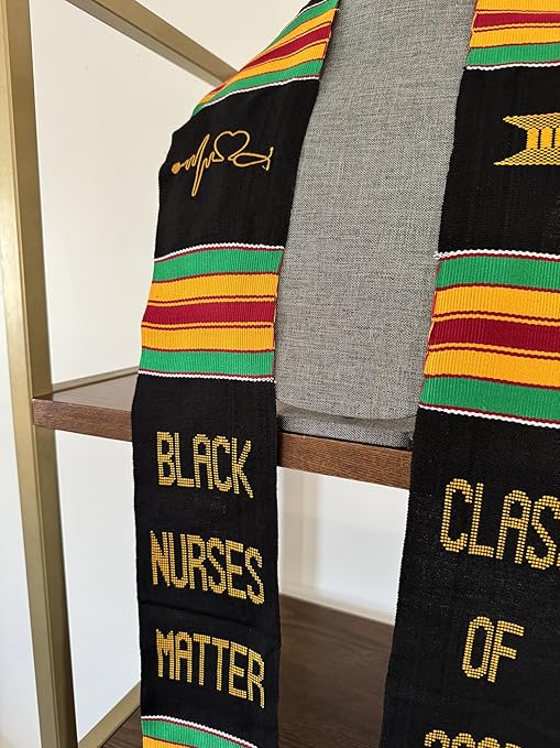 A black Kente graduation stole with vibrant red, yellow, and green stripes, draped over a gray mannequin. The stole features gold embroidery reading "BLACK NURSES MATTER" on one side and "CLASS OF 2025" on the other, along with a stethoscope heartbeat symbol and a gold pillar design.