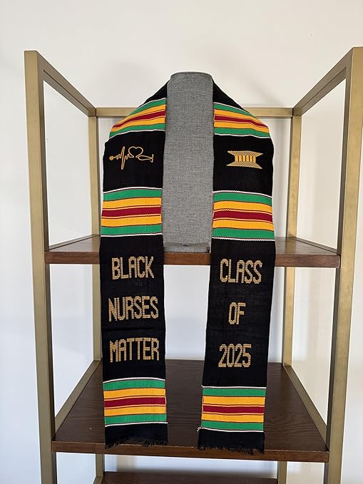 A black graduation stole with colorful Kente patterns and gold embroidery, designed to honor Black nurses. It features the phrase "BLACK NURSES MATTER" on the left panel and "CLASS OF 2025" on the right, with a heartbeat stethoscope symbol and a gold pillar icon.