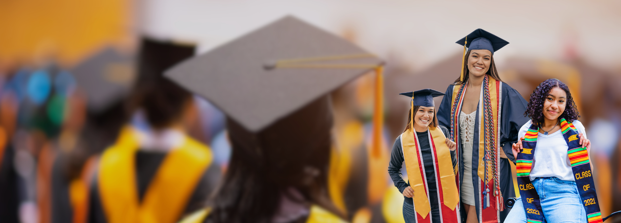 Graduates wearing caps and gowns with colorful stoles, including kente and gold designs, smiling and posing in the foreground, with a blurred background of a graduation ceremony showing other graduates in caps and gowns.