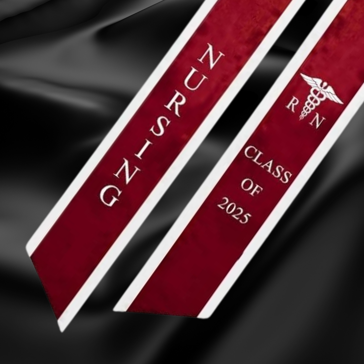 A burgundy nursing graduation stole with white trim, displaying bold vertical 'NURSING' lettering on the left, a medical logo with 'RN' at the top right, and 'CLASS OF 2025' embroidered horizontally below. The stole features slanted tips and a white plain background. All characters are embroidered. This photo is zoomed in with black satin cloth in the background.