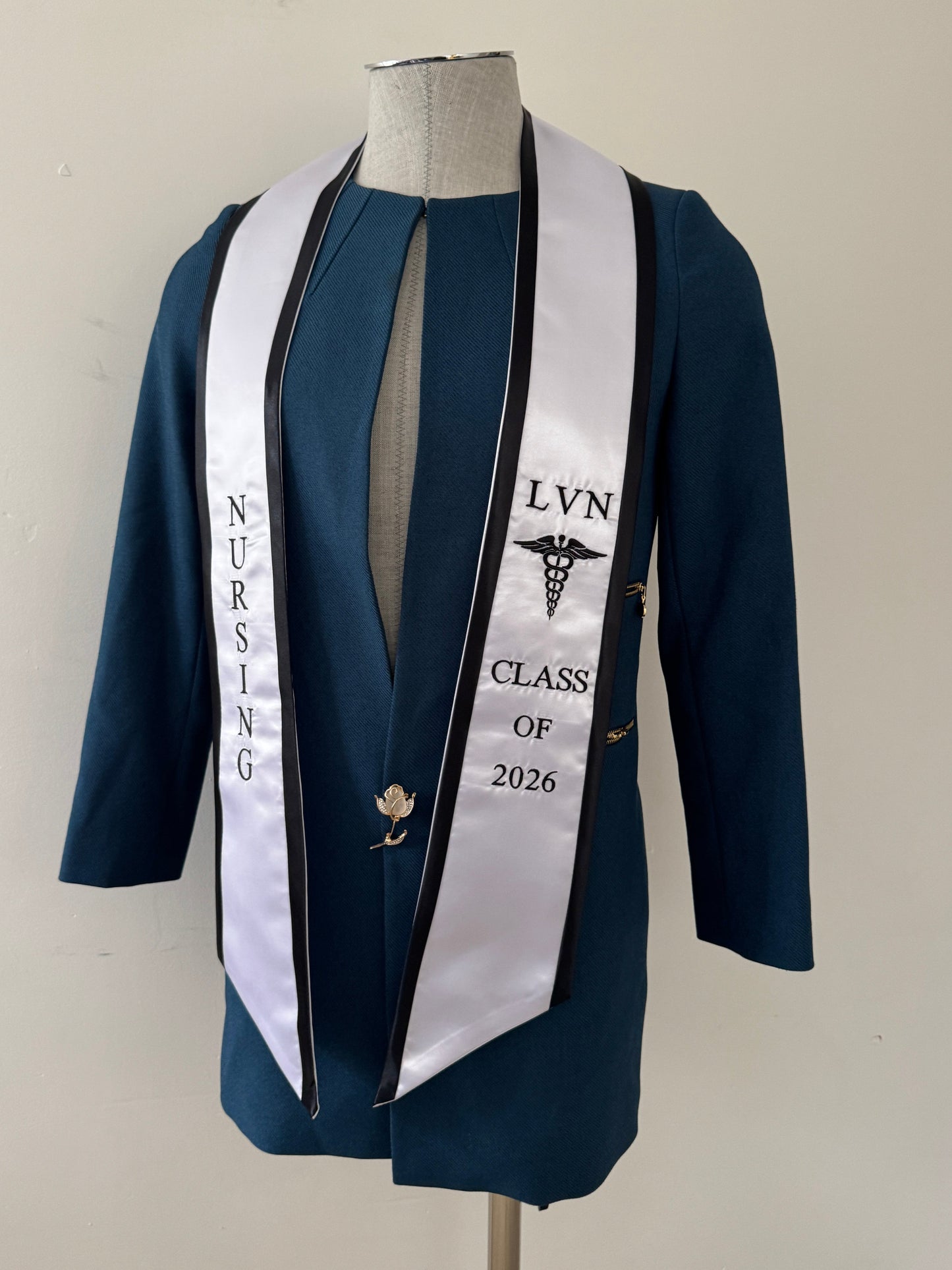 A White With Black Trim LVN graduation stole, showcasing bold 'LVN' lettering embroidered at the left, followed by a medical logo and 'CLASS OF 2026' embroidered beneath, and 'NURSING' lettering on the right. The stole has slanted tips and a white plain background. All characters are embroidered.