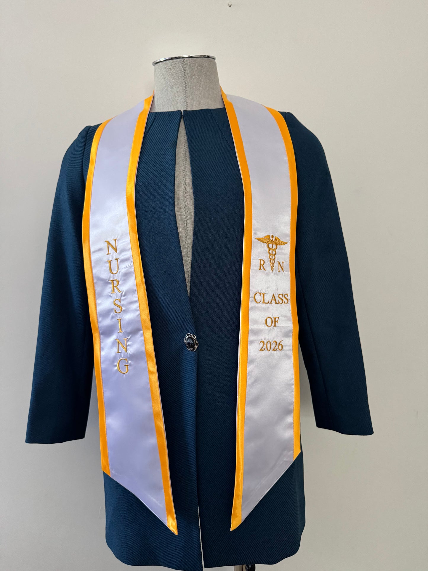 A White With Gold Trim RN Graduation Stole, displaying bold vertical 'NURSING' lettering on the right, a medical logo with 'RN' at the left, and 'CLASS OF 2026' embroidered horizontally below. The stole features slanted tips and a white plain background. All characters are embroidered.