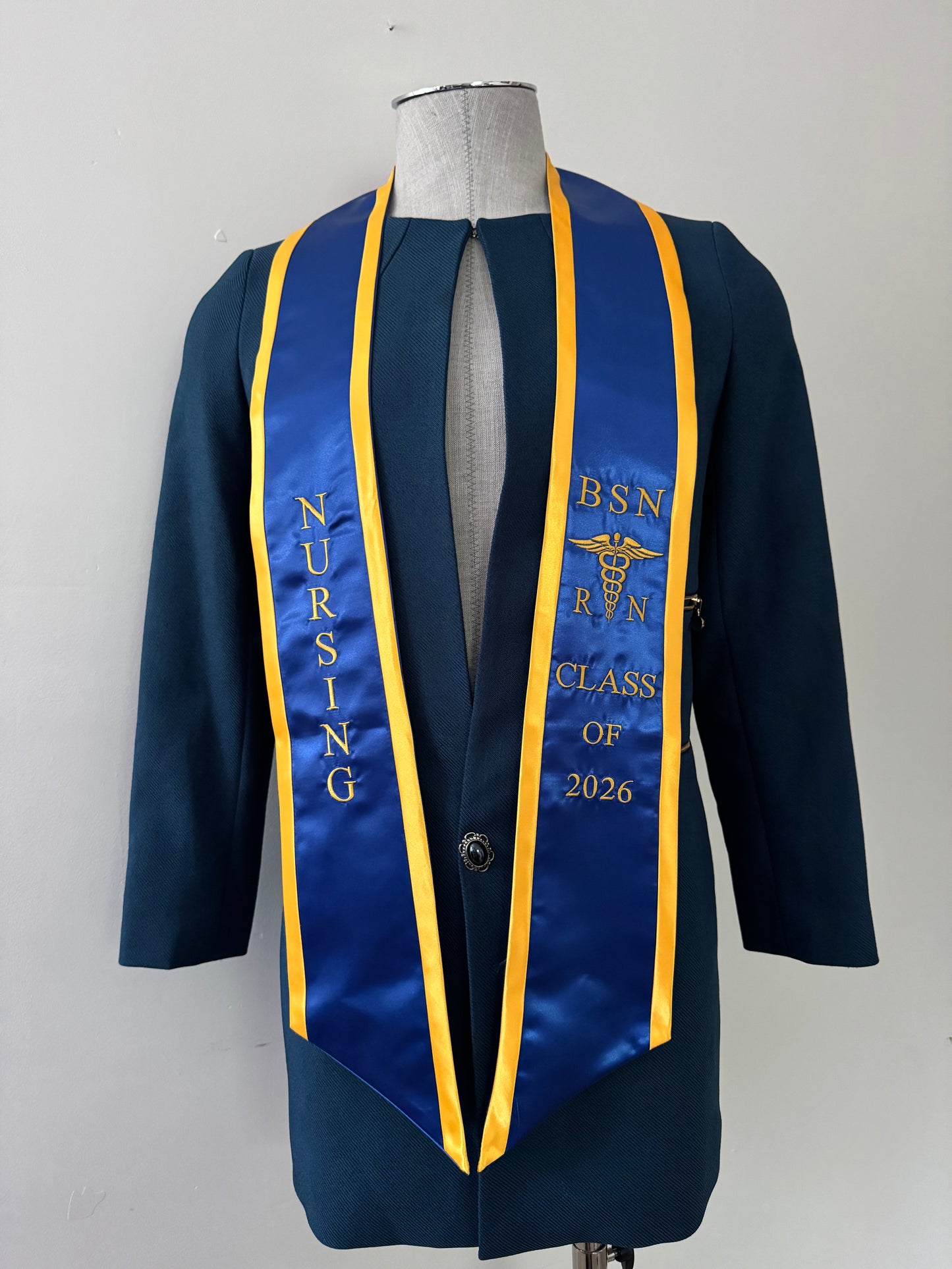 A blue with gold trim BSN RN graduation stole, displaying bold 'BSN RN' lettering embroidered on the left, a medical logo with BSN RN on the right, and 'CLASS OF 2026' embroidered horizontally below. The stole has slanted tips and a white plain background. All text is embroidered.