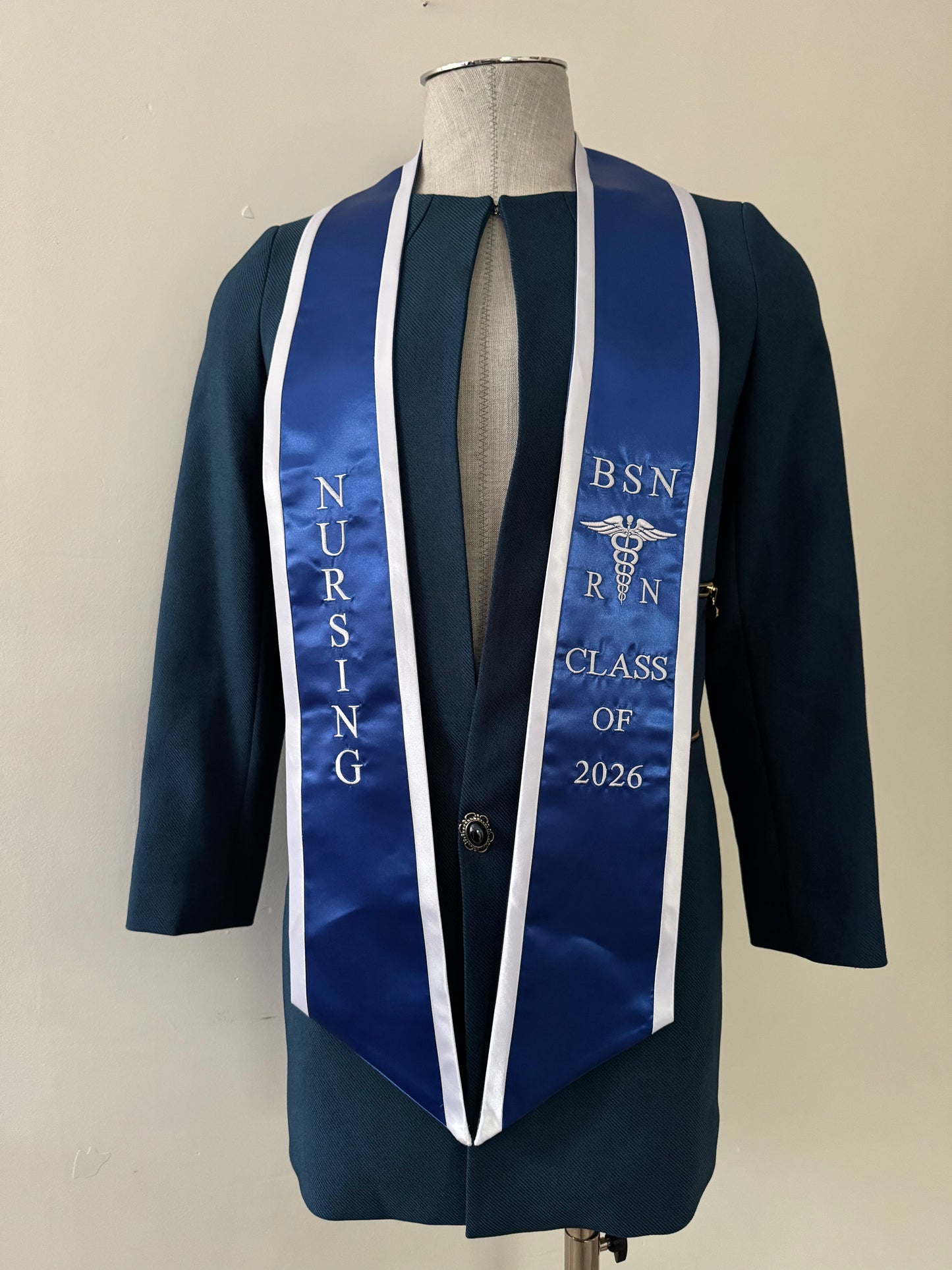 A blue BSN RN graduation stole with white trim, displaying bold 'BSN RN' lettering embroidered on the left, a medical logo with BSN RN on the right, and 'CLASS OF 2026' embroidered horizontally below. The stole has slanted tips and a white plain background. All text is embroidered.