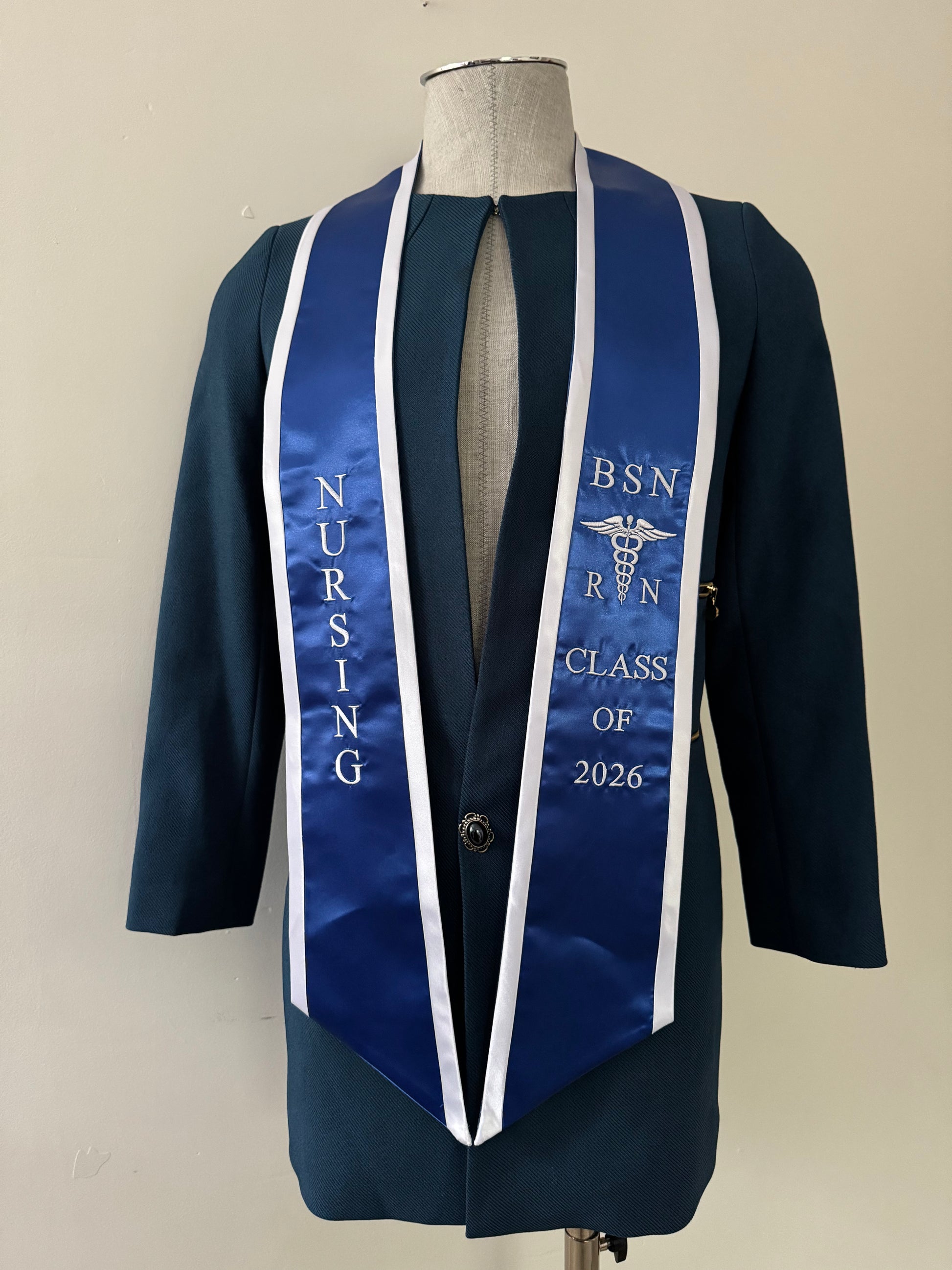 A blue BSN RN graduation stole with white trim, displaying bold 'BSN RN' lettering embroidered on the left, a medical logo with BSN RN on the right, and 'CLASS OF 2026' embroidered horizontally below. The stole has slanted tips and a white plain background. All text is embroidered.