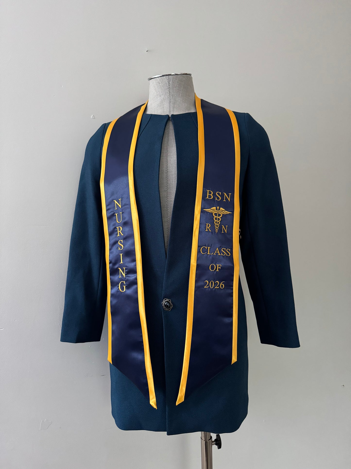 A navy blue with gold trim BSN RN graduation stole, displaying bold 'BSN RN' lettering embroidered on the left, a medical logo with BSN RN on the right, and 'CLASS OF 2026' embroidered horizontally below. The stole has slanted tips and a white plain background. All text is embroidered.