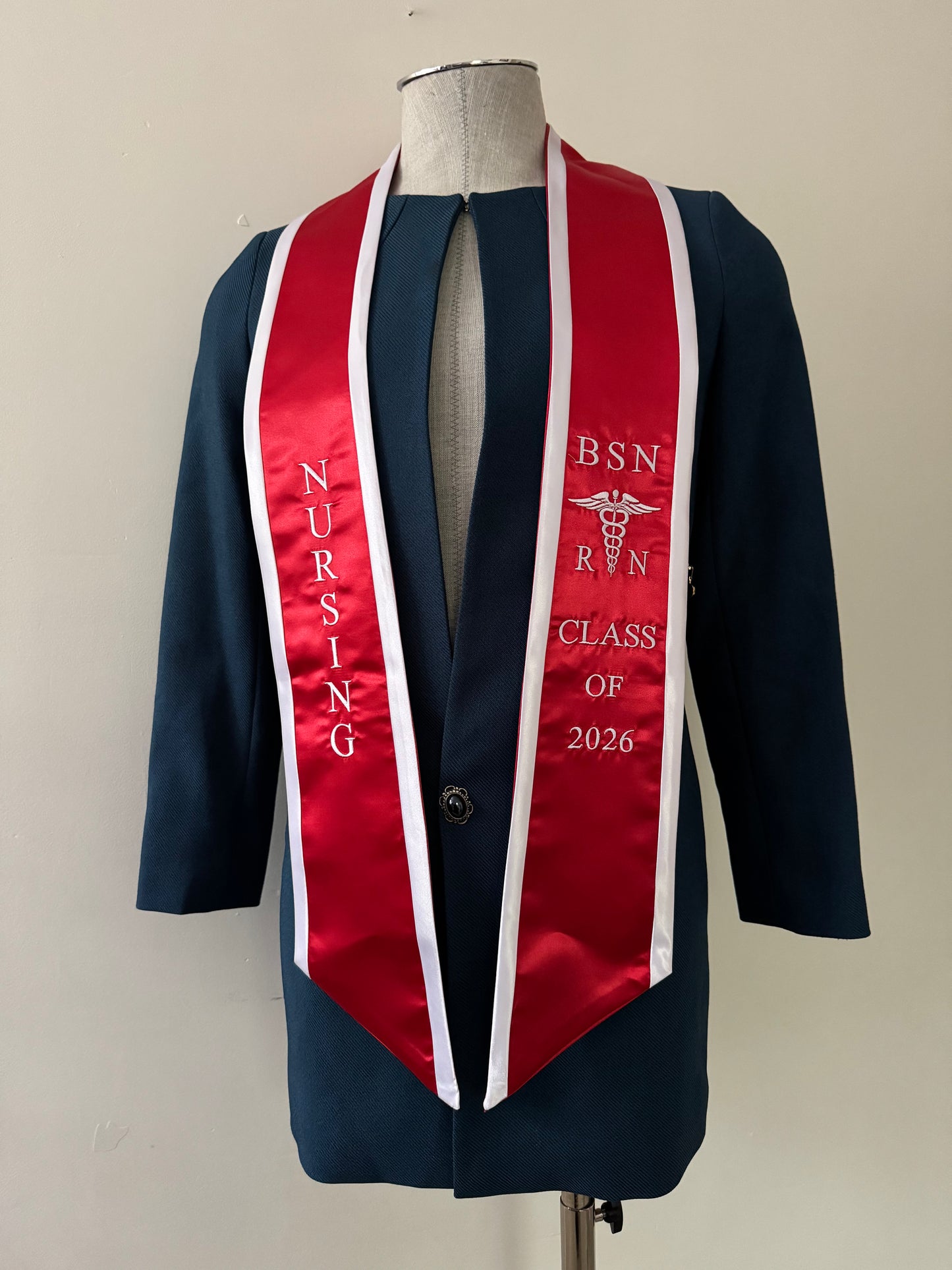 A red with white trim BSN RN graduation stole, displaying bold 'BSN RN' lettering embroidered on the left, a medical logo with BSN RN on the right, and 'CLASS OF 2026' embroidered horizontally below. The stole has slanted tips and a white plain background. All text is embroidered.