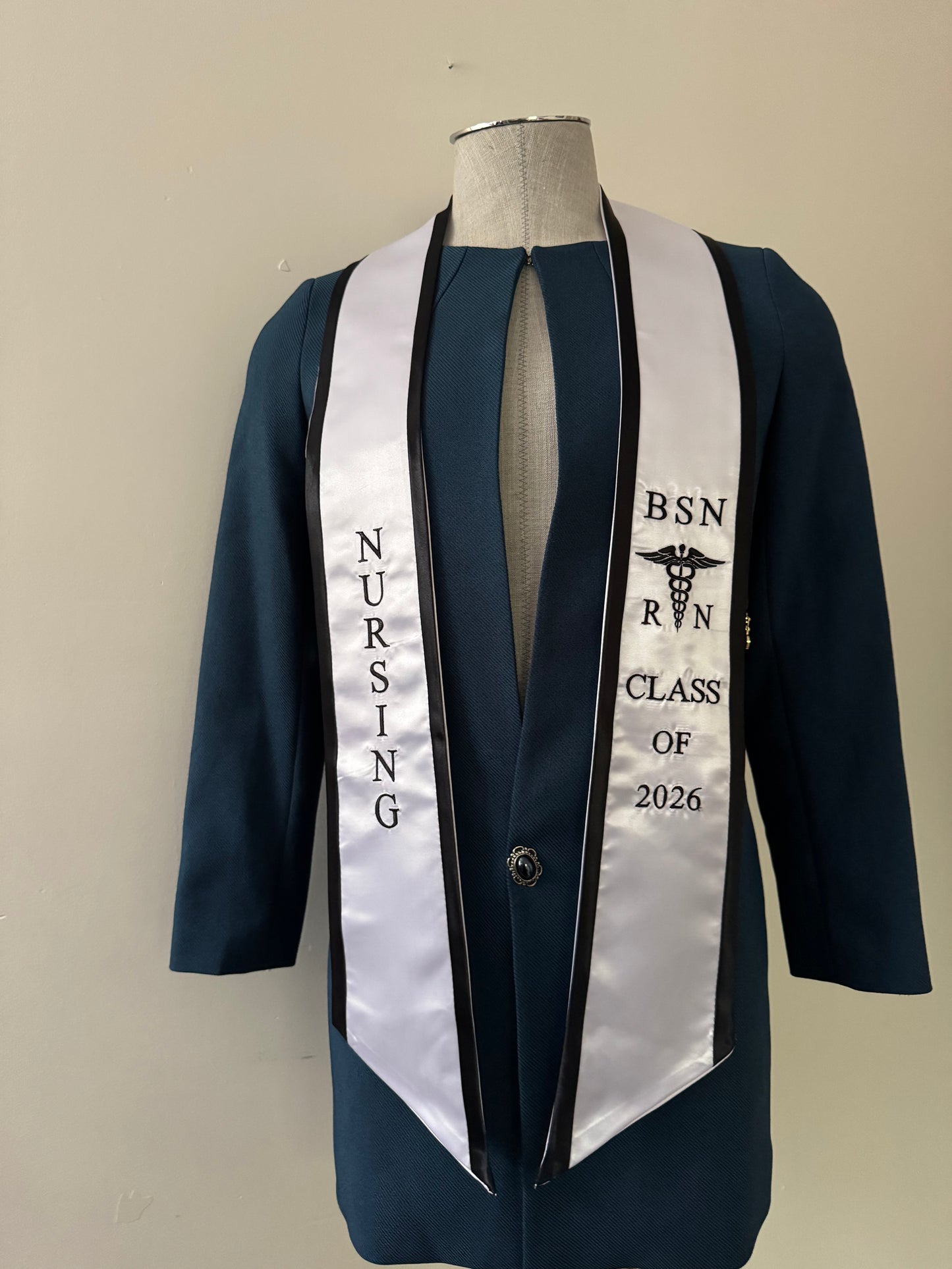 A white with black trim BSN RN graduation stole, displaying bold 'BSN RN' lettering embroidered on the left, a medical logo with BSN RN on the right, and 'CLASS OF 2026' embroidered horizontally below. The stole has slanted tips and a white plain background. All text is embroidered.