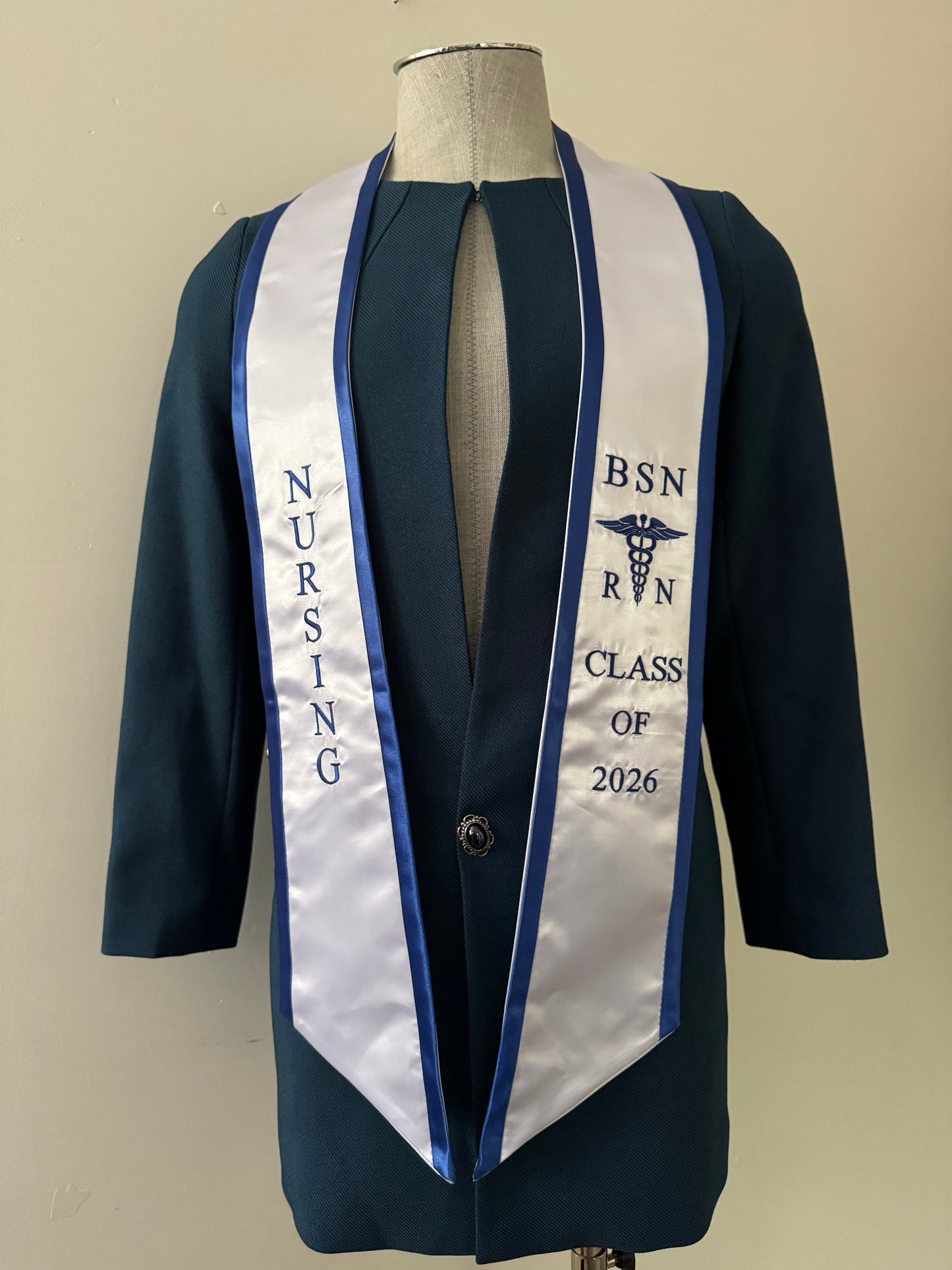 A white BSN RN graduation stole with blue trim, displaying bold 'BSN RN' lettering embroidered on the left, a medical logo with BSN RN on the right, and 'CLASS OF 2026' embroidered horizontally below. The stole has slanted tips and a white plain background. All text is embroidered.