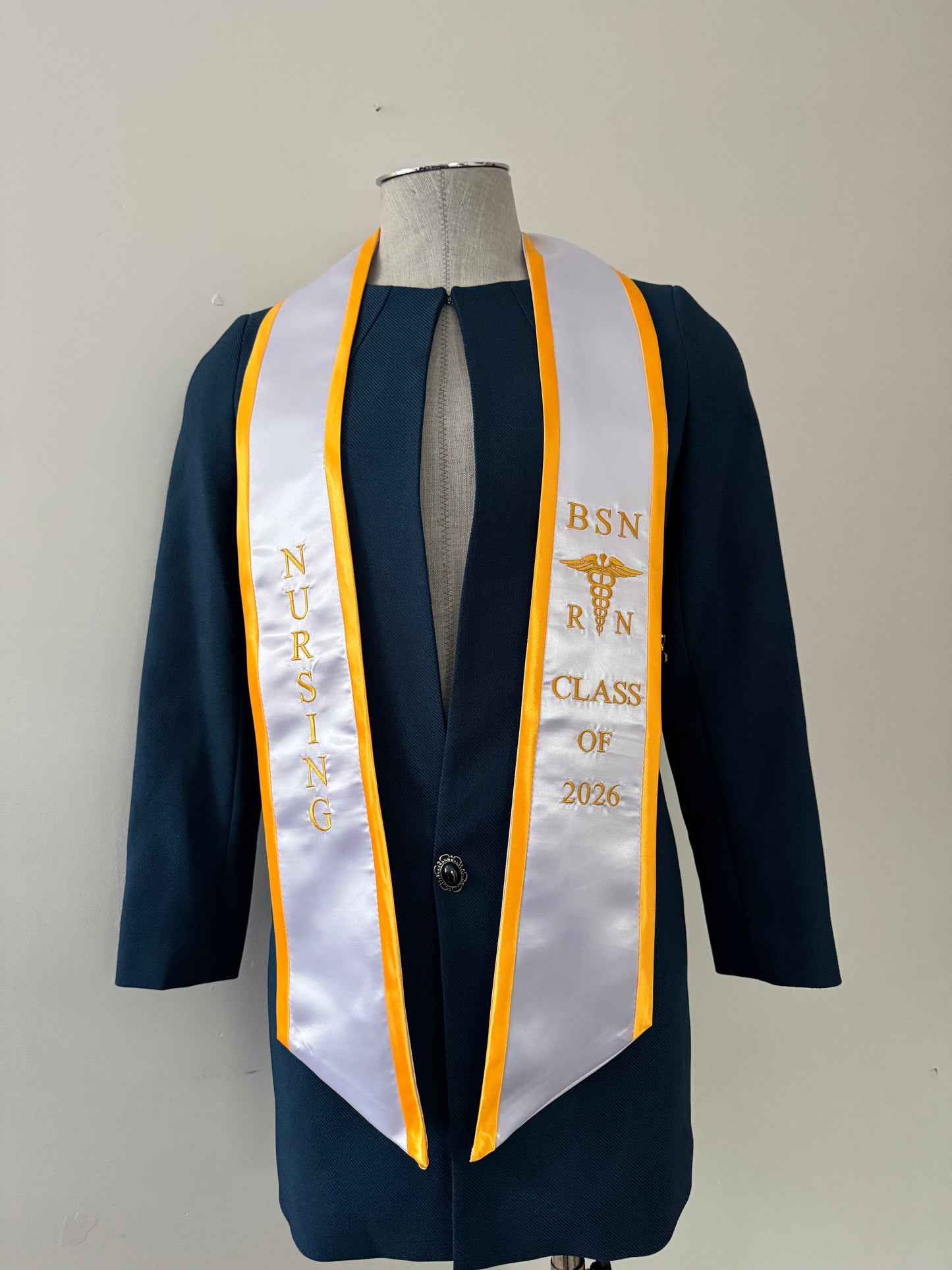 A white with gold trim BSN RN graduation stole, displaying bold 'BSN RN' lettering embroidered on the left, a medical logo with BSN RN on the right, and 'CLASS OF 2026' embroidered horizontally below. The stole has slanted tips and a white plain background. All text is embroidered.