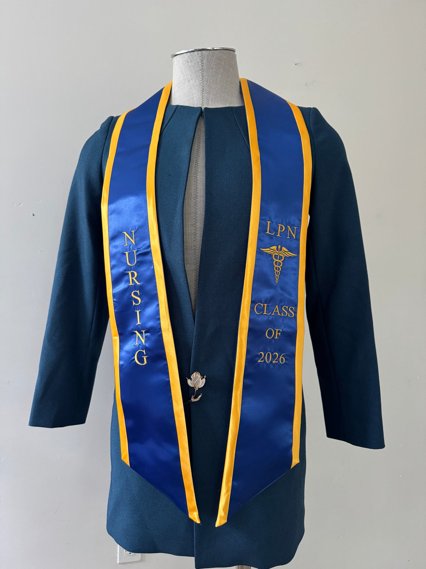 A Blue With Gold Trim LPN graduation stole with royal blue trim, showcasing bold 'LPN ' lettering embroidered at the top, followed by a medical logo and 'CLASS OF 2026' embroidered beneath. The stole has slanted tips and a white plain background. All characters are embroidered.