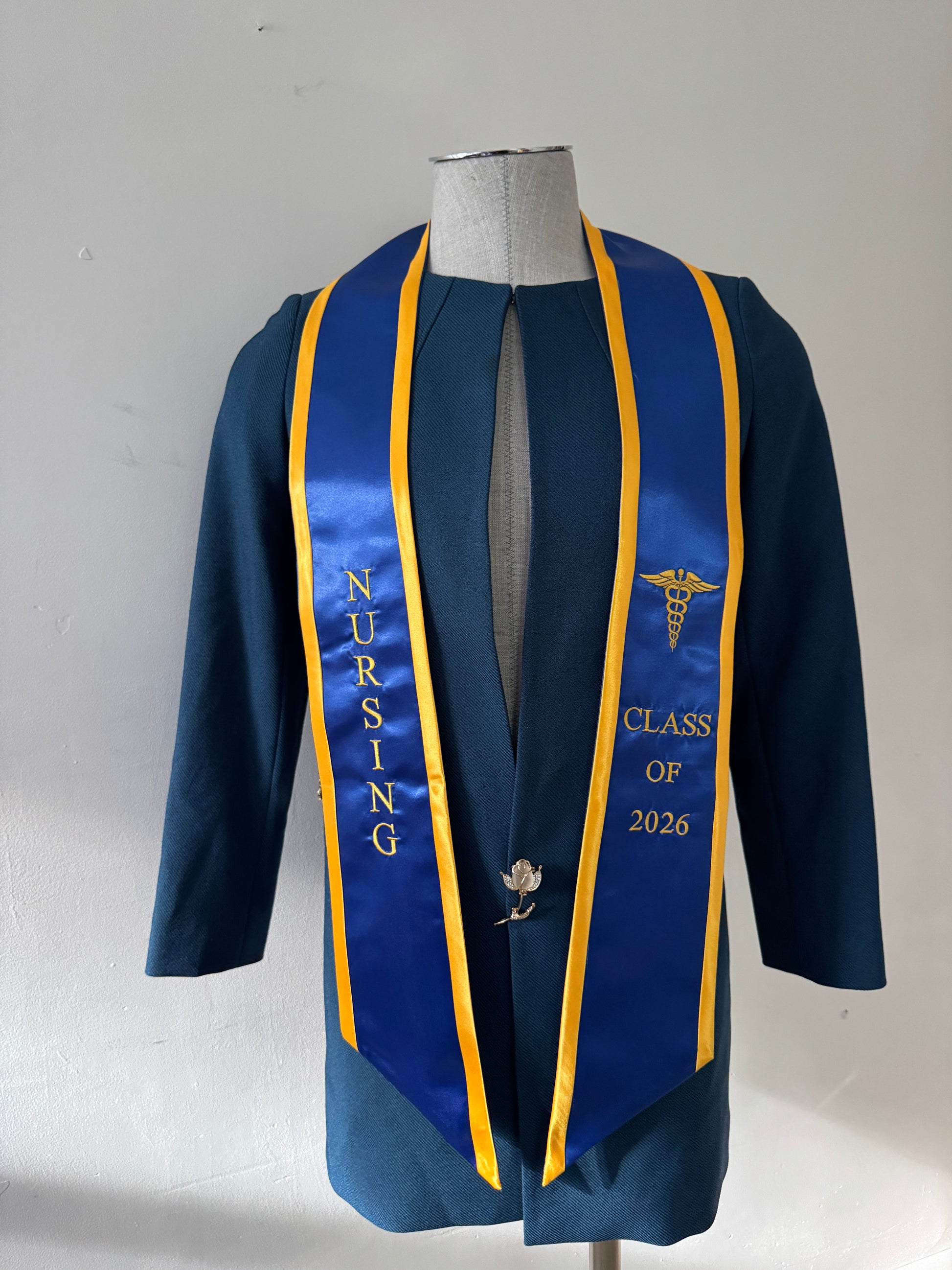 A Blue With Gold Trim Nursing Graduation Stole, displaying bold 'CLASS OF 2026' lettering embroidered on the left, a medical logo with NURSING on the right embroidered horizontally below. The stole has slanted tips and a white plain background. All text is embroidered.