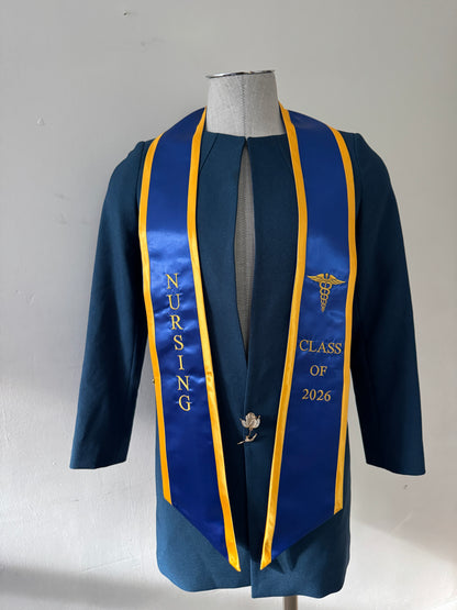 A Blue With Gold Trim Nursing Graduation Stole, displaying bold 'CLASS OF 2026' lettering embroidered on the left, a medical logo with NURSING on the right embroidered horizontally below. The stole has slanted tips and a white plain background. All text is embroidered.