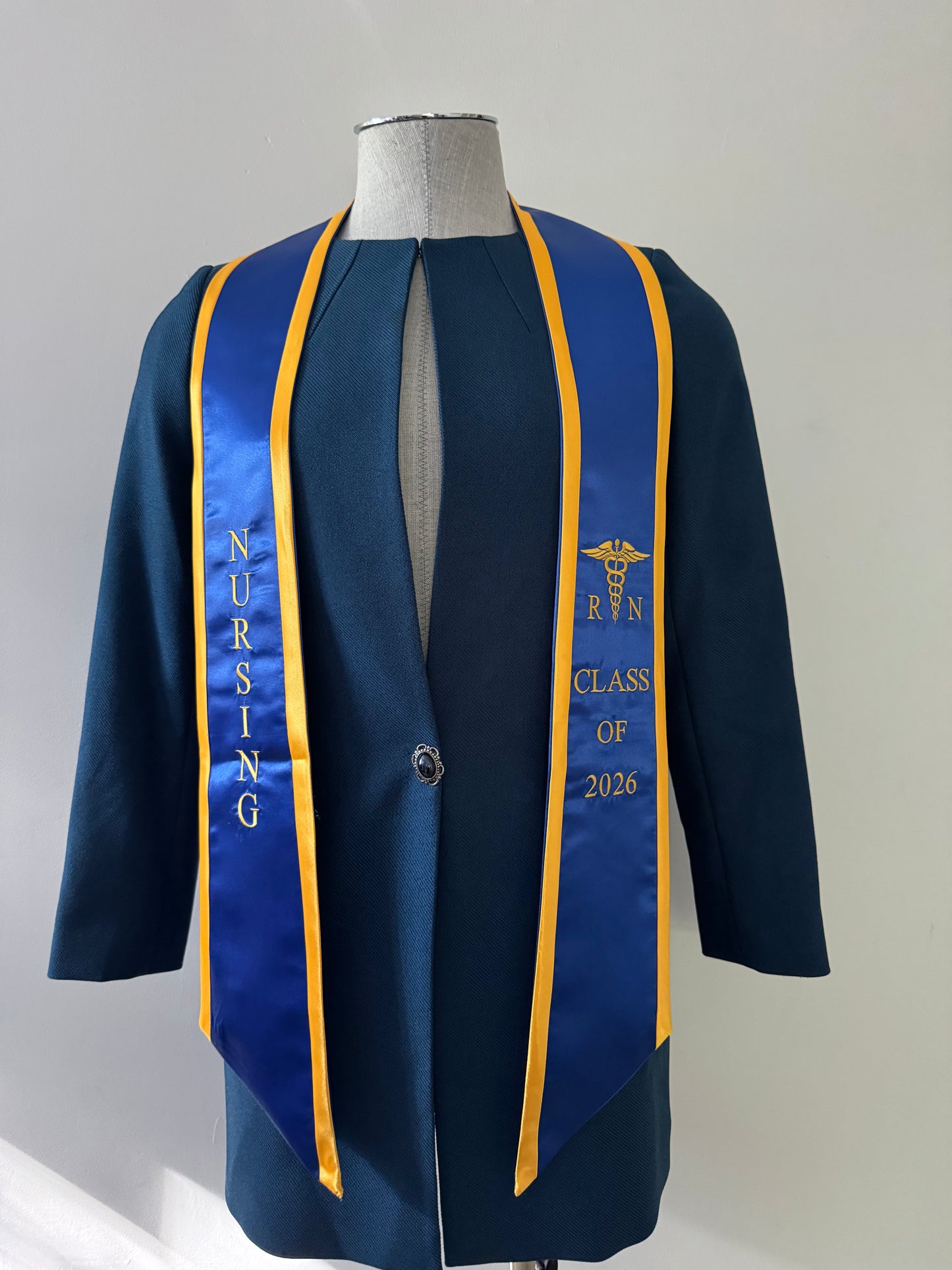 A Blue With Gold Trim RN Graduation Stole, displaying bold vertical 'NURSING' lettering on the right, a medical logo with 'RN' at the left, and 'CLASS OF 2026' embroidered horizontally below. The stole features slanted tips and a white plain background. All characters are embroidered.