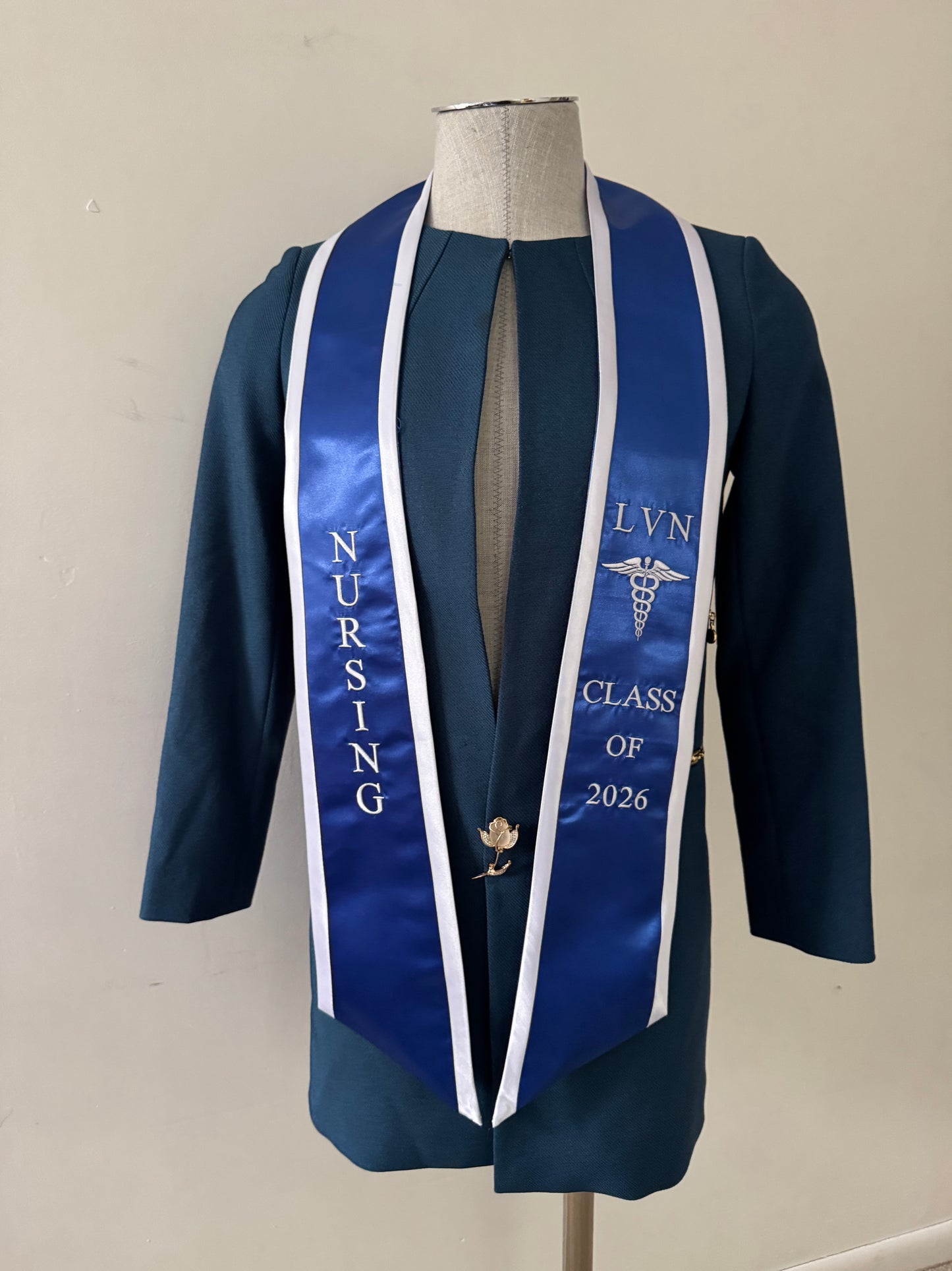 A Blue With White Trim LVN graduation stole, showcasing bold 'LVN' lettering embroidered at the left, followed by a medical logo and 'CLASS OF 2026' embroidered beneath, and 'NURSING' lettering on the right. The stole has slanted tips and a white plain background. All characters are embroidered.
