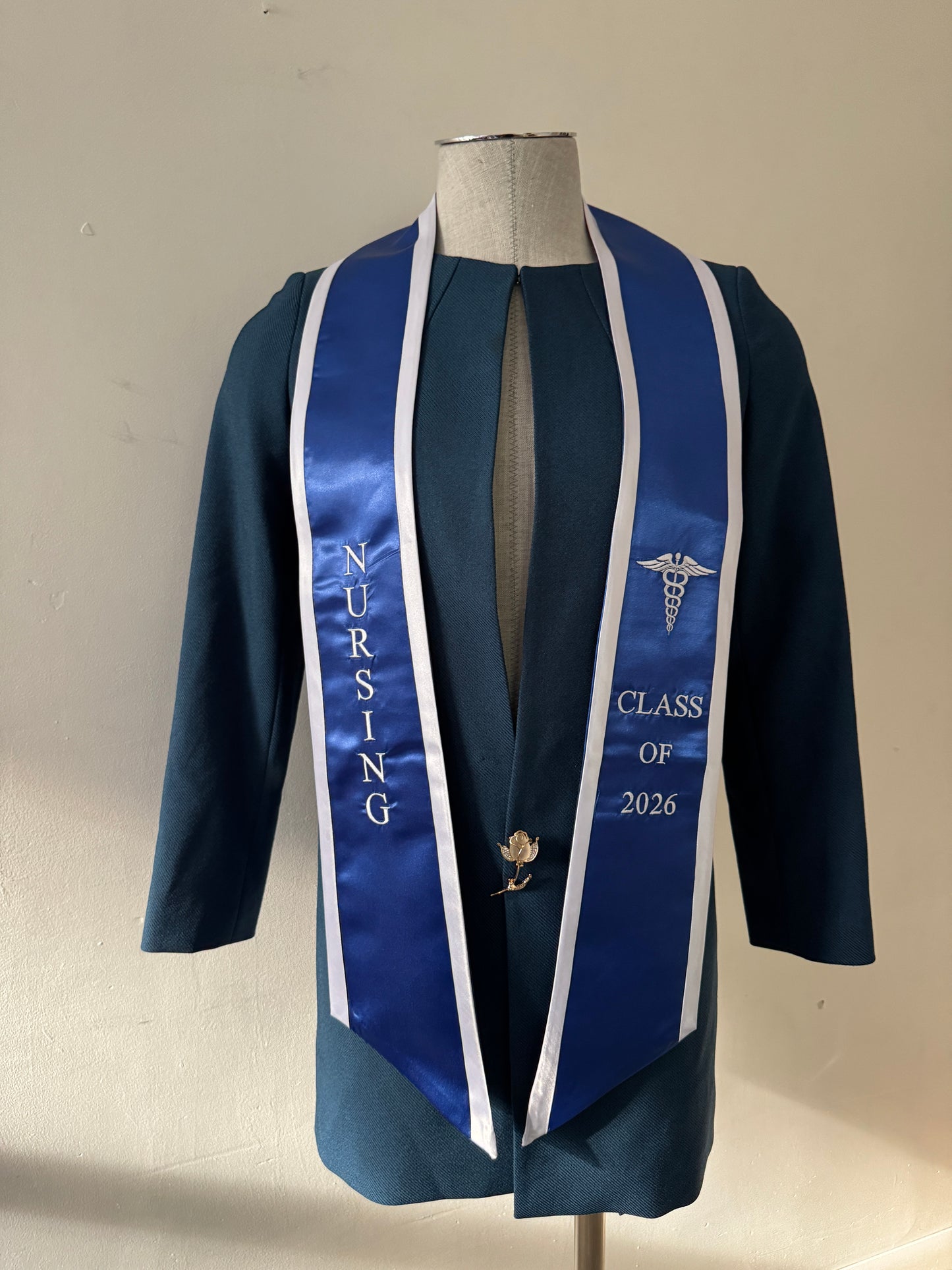 A Blue With White Trim Nursing Graduation Stole, displaying bold 'CLASS OF 2026' lettering embroidered on the left, a medical logo with NURSING on the right embroidered horizontally below. The stole has slanted tips and a white plain background. All text is embroidered.