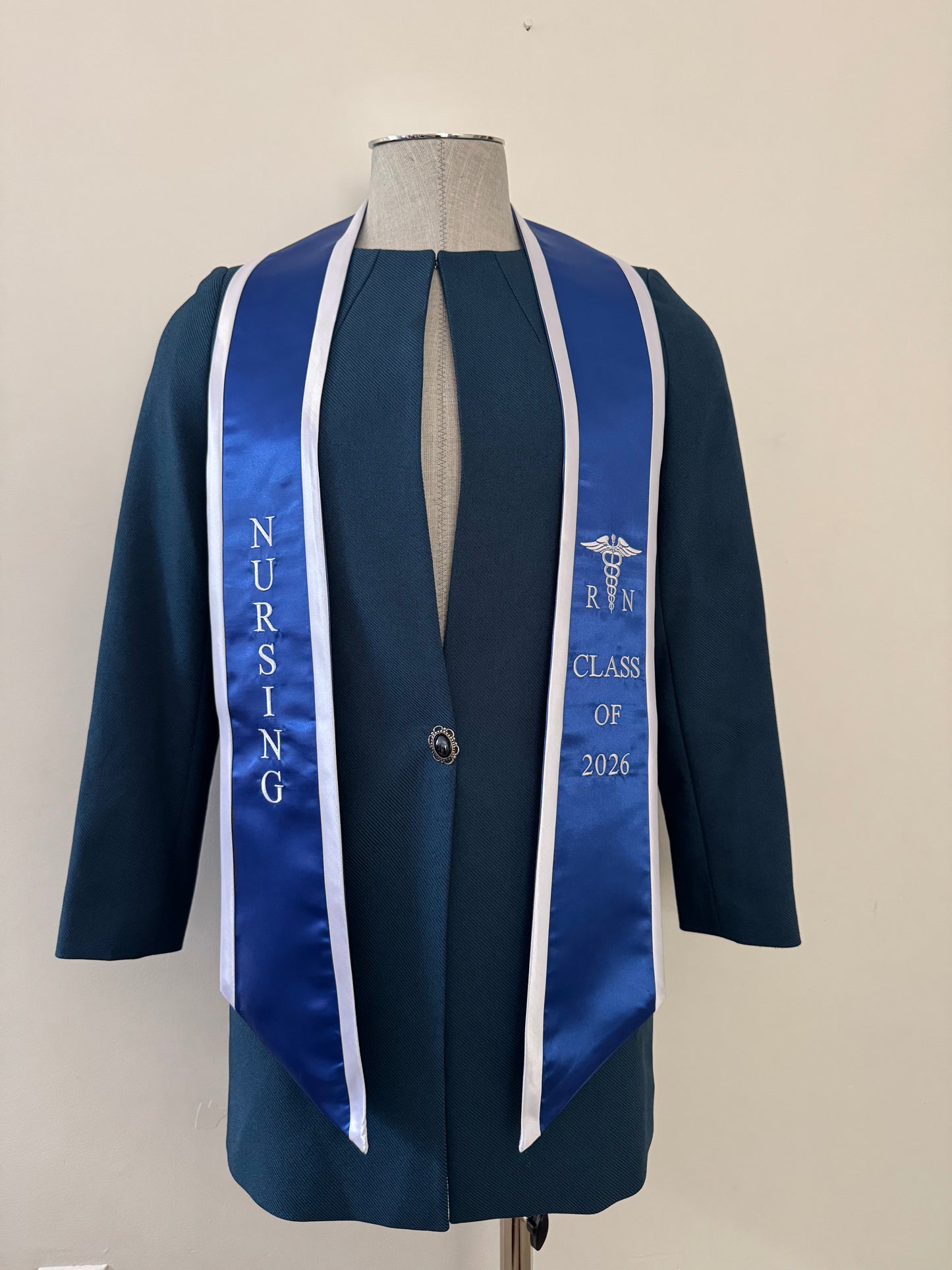 A Blue With White Trim RN Graduation Stole, displaying bold vertical 'NURSING' lettering on the right, a medical logo with 'RN' at the left, and 'CLASS OF 2026' embroidered horizontally below. The stole features slanted tips and a white plain background. All characters are embroidered.