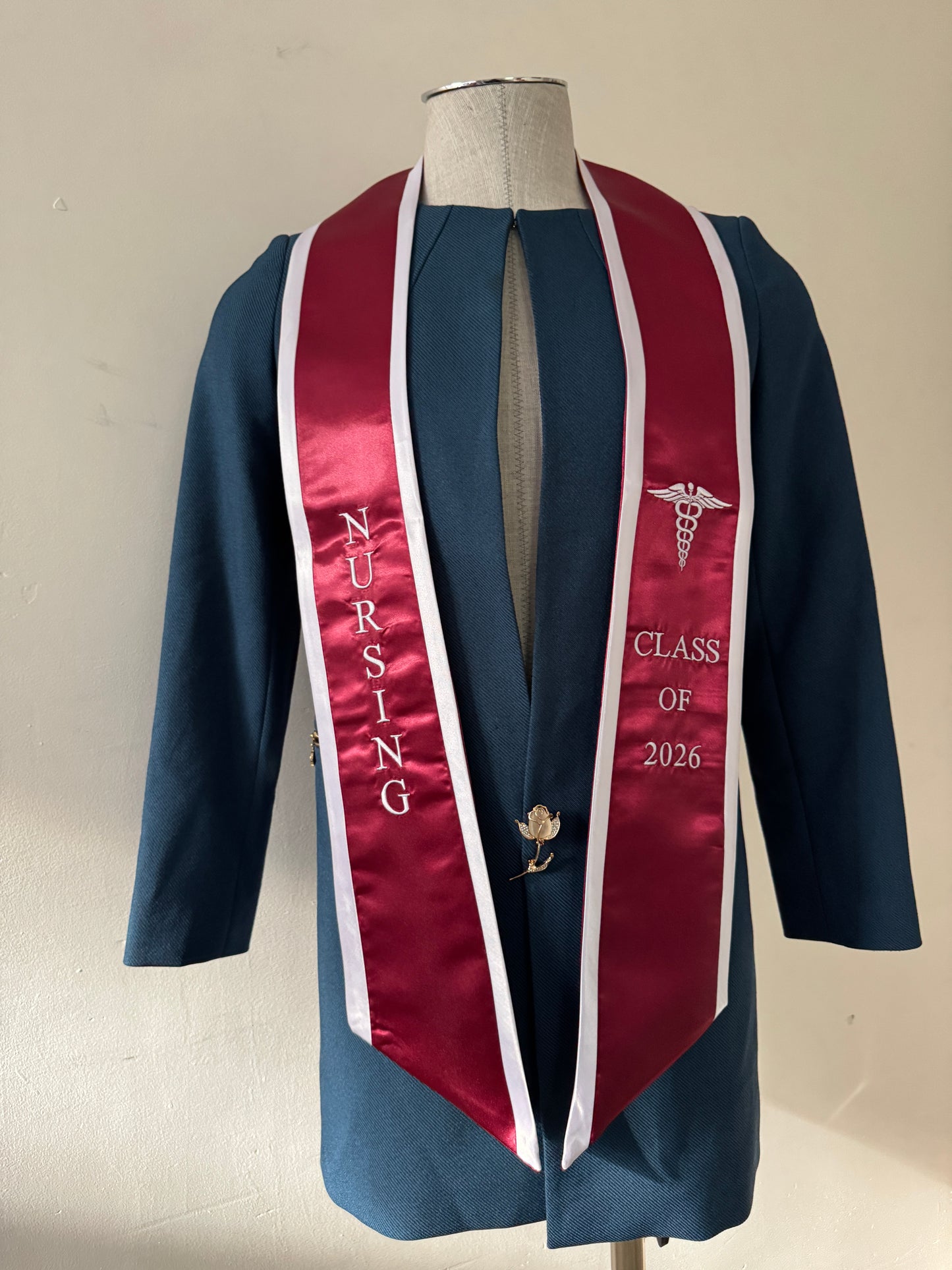 A Burgundy With White Nursing Graduation Stole, displaying bold 'CLASS OF 2026' lettering embroidered on the left, a medical logo with NURSING on the right embroidered horizontally below. The stole has slanted tips and a white plain background. All text is embroidered.