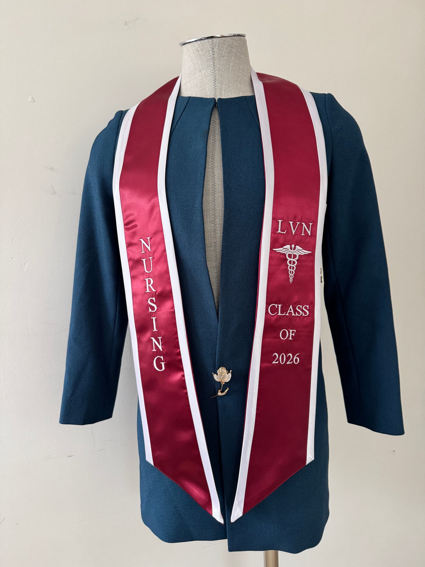 A Burgundy With White Trim LVN graduation stole, showcasing bold 'LVN' lettering embroidered at the left, followed by a medical logo and 'CLASS OF 2026' embroidered beneath, and 'NURSING' lettering on the right. The stole has slanted tips and a white plain background. All characters are embroidered.