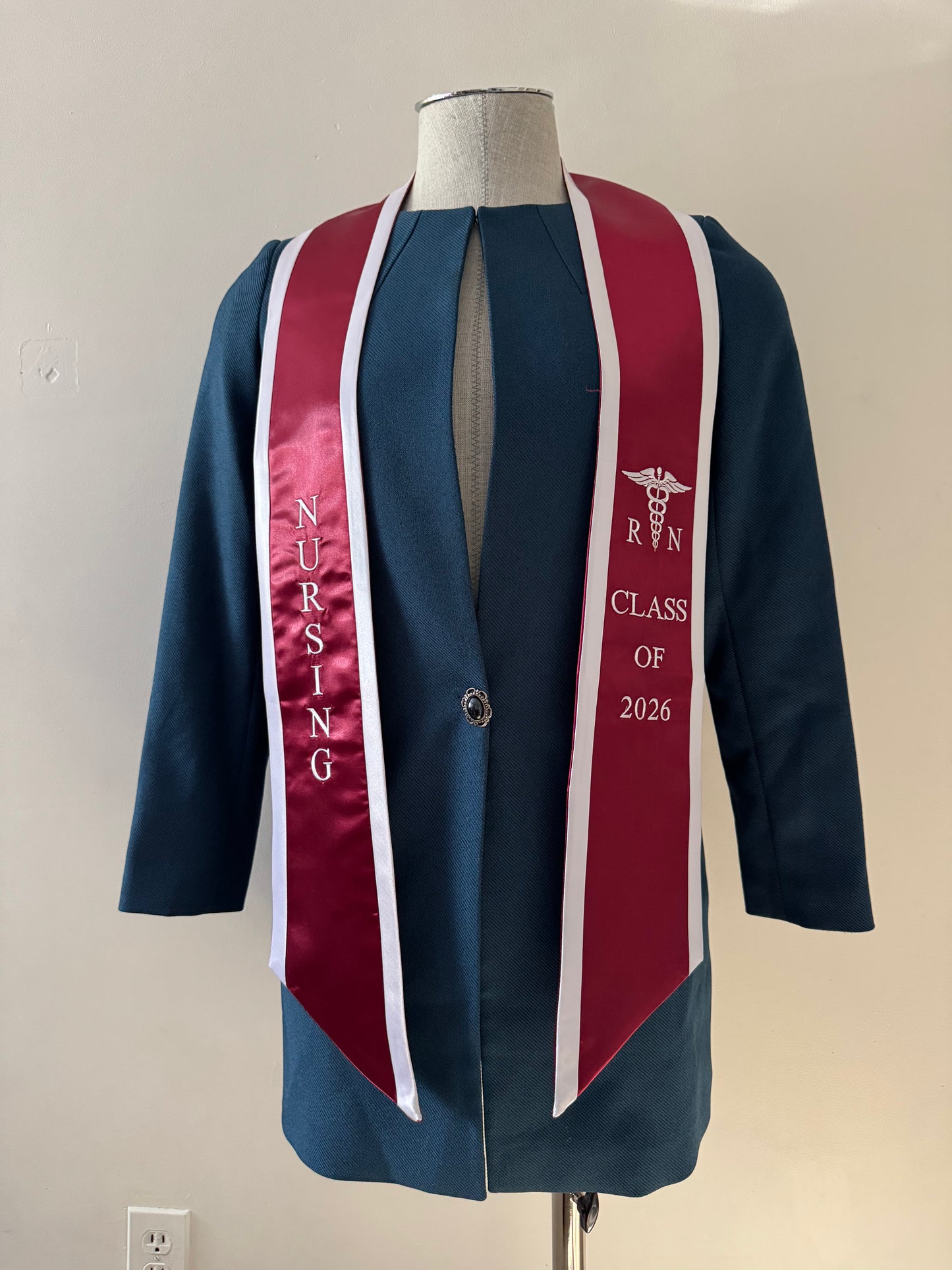 A Burgundy With White Trim RN Graduation Stole, displaying bold vertical 'NURSING' lettering on the right, a medical logo with 'RN' at the left, and 'CLASS OF 2026' embroidered horizontally below. The stole features slanted tips and a white plain background. All characters are embroidered.