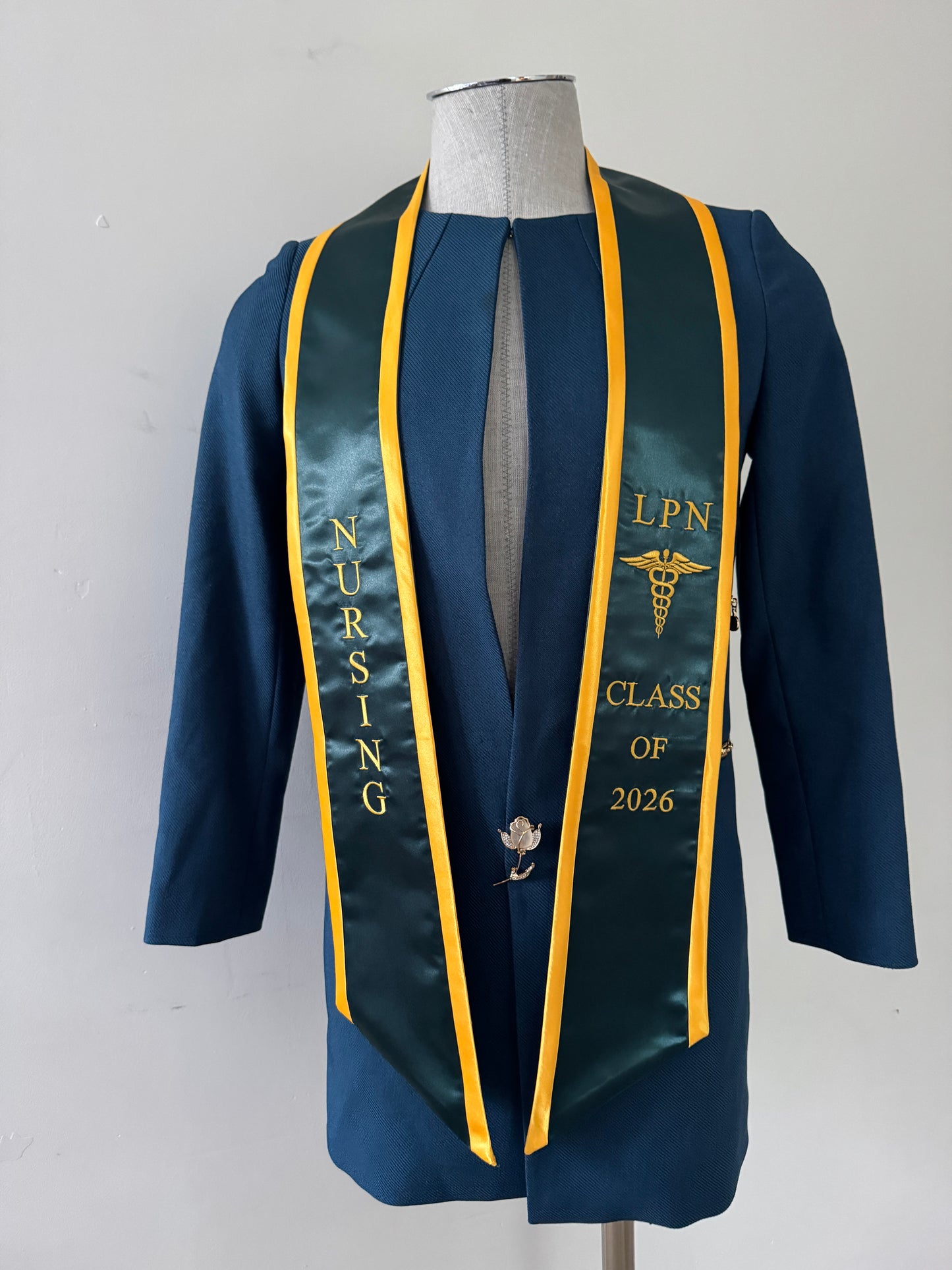 A Forest Green With Gold Trim LPN graduation stole with royal blue trim, showcasing bold 'LPN ' lettering embroidered at the top, followed by a medical logo and 'CLASS OF 2026' embroidered beneath. The stole has slanted tips and a white plain background. All characters are embroidered.