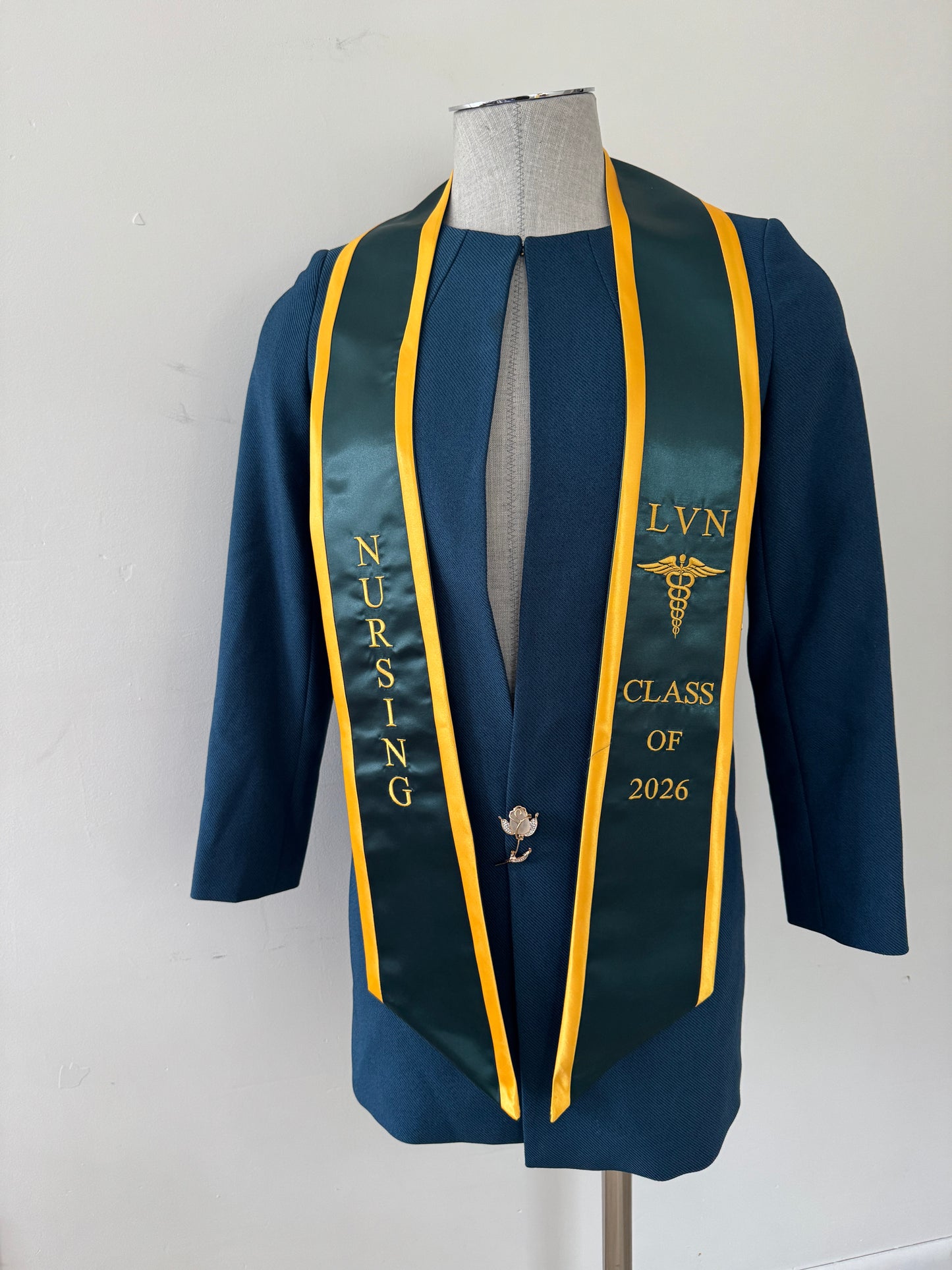 A Forest Green With Gold Trim LVN graduation stole, showcasing bold 'LVN' lettering embroidered at the left, followed by a medical logo and 'CLASS OF 2026' embroidered beneath, and 'NURSING' lettering on the right. The stole has slanted tips and a white plain background. All characters are embroidered.