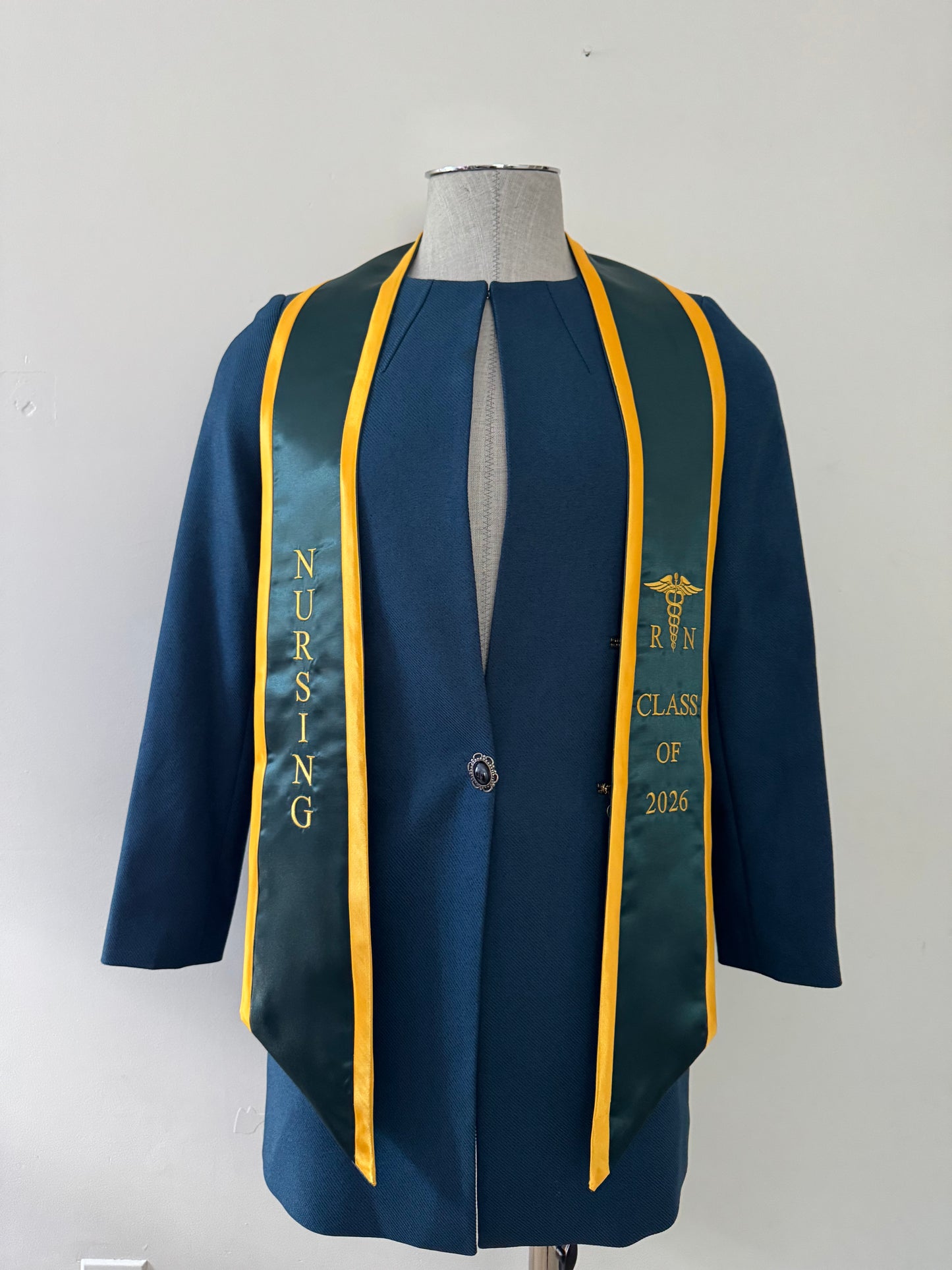A Forest Green With Gold Trim RN Graduation Stole, displaying bold vertical 'NURSING' lettering on the right, a medical logo with 'RN' at the left, and 'CLASS OF 2026' embroidered horizontally below. The stole features slanted tips and a white plain background. All characters are embroidered.