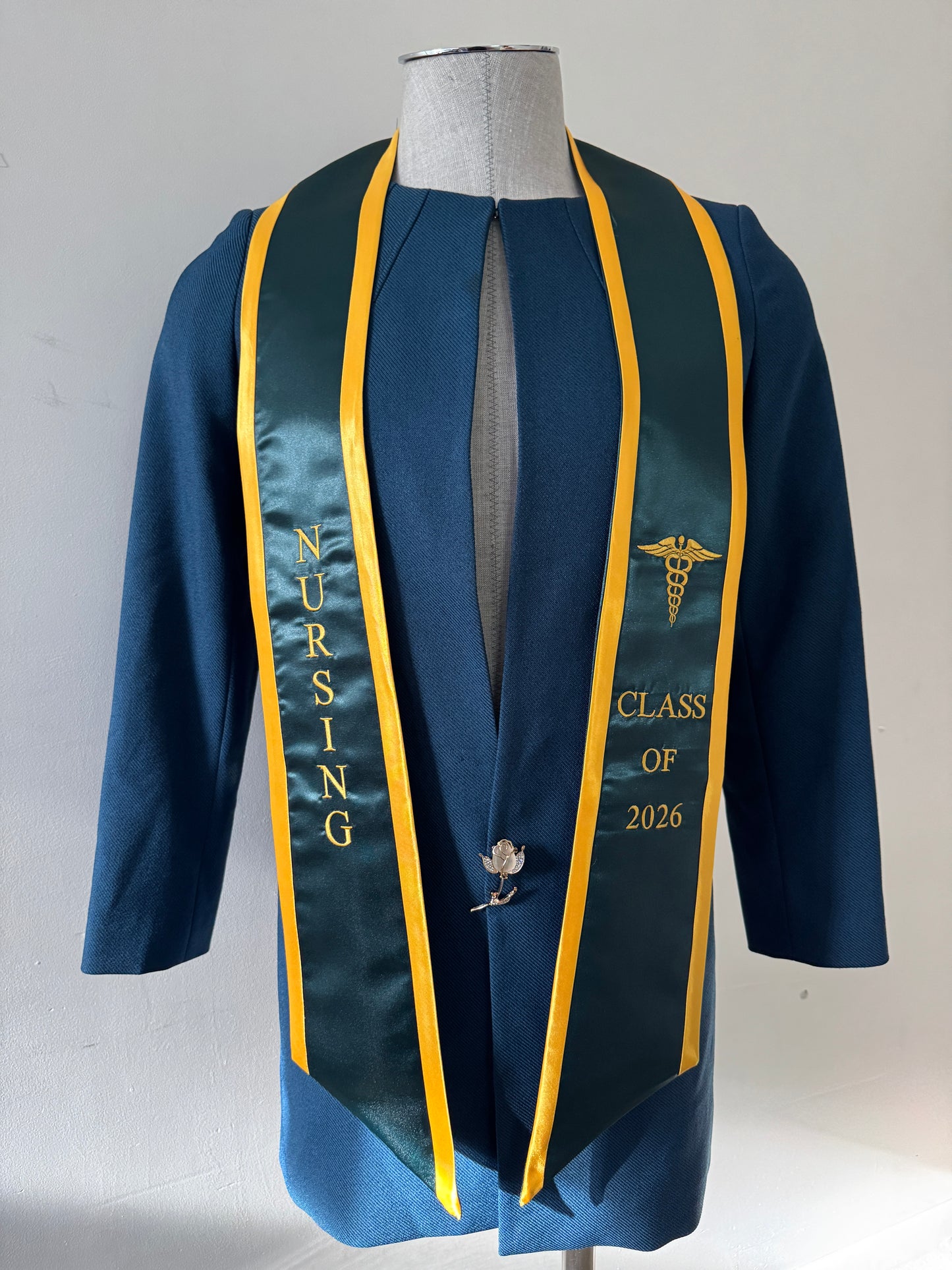 A Forest Green with Gold Trim Nursing Graduation Stole, displaying bold 'CLASS OF 2026' lettering embroidered on the left, a medical logo with NURSING on the right embroidered horizontally below. The stole has slanted tips and a white plain background. All text is embroidered.