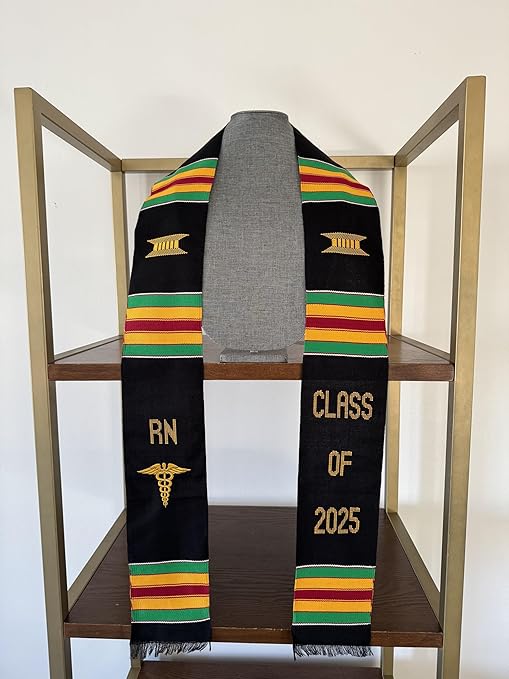 A black Kente graduation stole with vibrant red, yellow, and green stripes, draped over a gray mannequin. The stole features gold embroidery reading "RN" on one side and "CLASS OF 2025" on the other, along with a stethoscope heartbeat symbol and a gold pillar design.