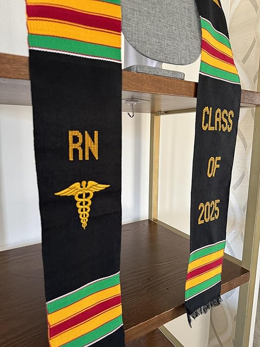 A black Kente graduation stole with vibrant red, yellow, and green stripes, draped over a gray mannequin. The stole features gold embroidery reading "RN" on one side and "CLASS OF 2025" on the other, along with a stethoscope heartbeat symbol and a gold pillar design.