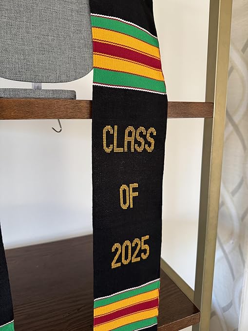 A black Kente graduation stole with vibrant red, yellow, and green stripes, draped over a gray mannequin. The stole features gold embroidery reading "RN" on one side and "CLASS OF 2025" on the other, along with a stethoscope heartbeat symbol and a gold pillar design.