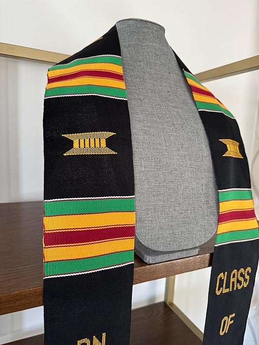 A black Kente graduation stole with vibrant red, yellow, and green stripes, draped over a gray mannequin. The stole features gold embroidery reading "RN" on one side and "CLASS OF 2025" on the other, along with a stethoscope heartbeat symbol and a gold pillar design.