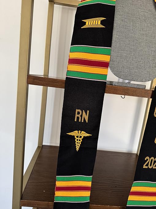 A black Kente graduation stole with vibrant red, yellow, and green stripes, draped over a gray mannequin. The stole features gold embroidery reading "RN" on one side and "CLASS OF 2025" on the other, along with a stethoscope heartbeat symbol and a gold pillar design.