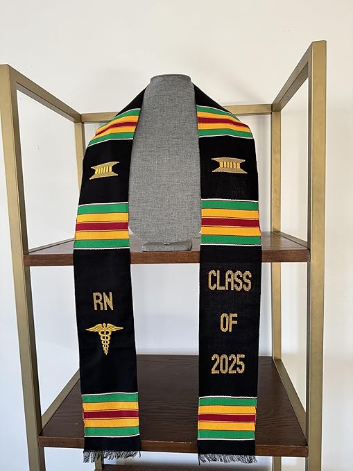 A black Kente graduation stole with vibrant red, yellow, and green stripes, draped over a gray mannequin. The stole features gold embroidery reading "RN" on one side and "CLASS OF 2025" on the other, along with a stethoscope heartbeat symbol and a gold pillar design.