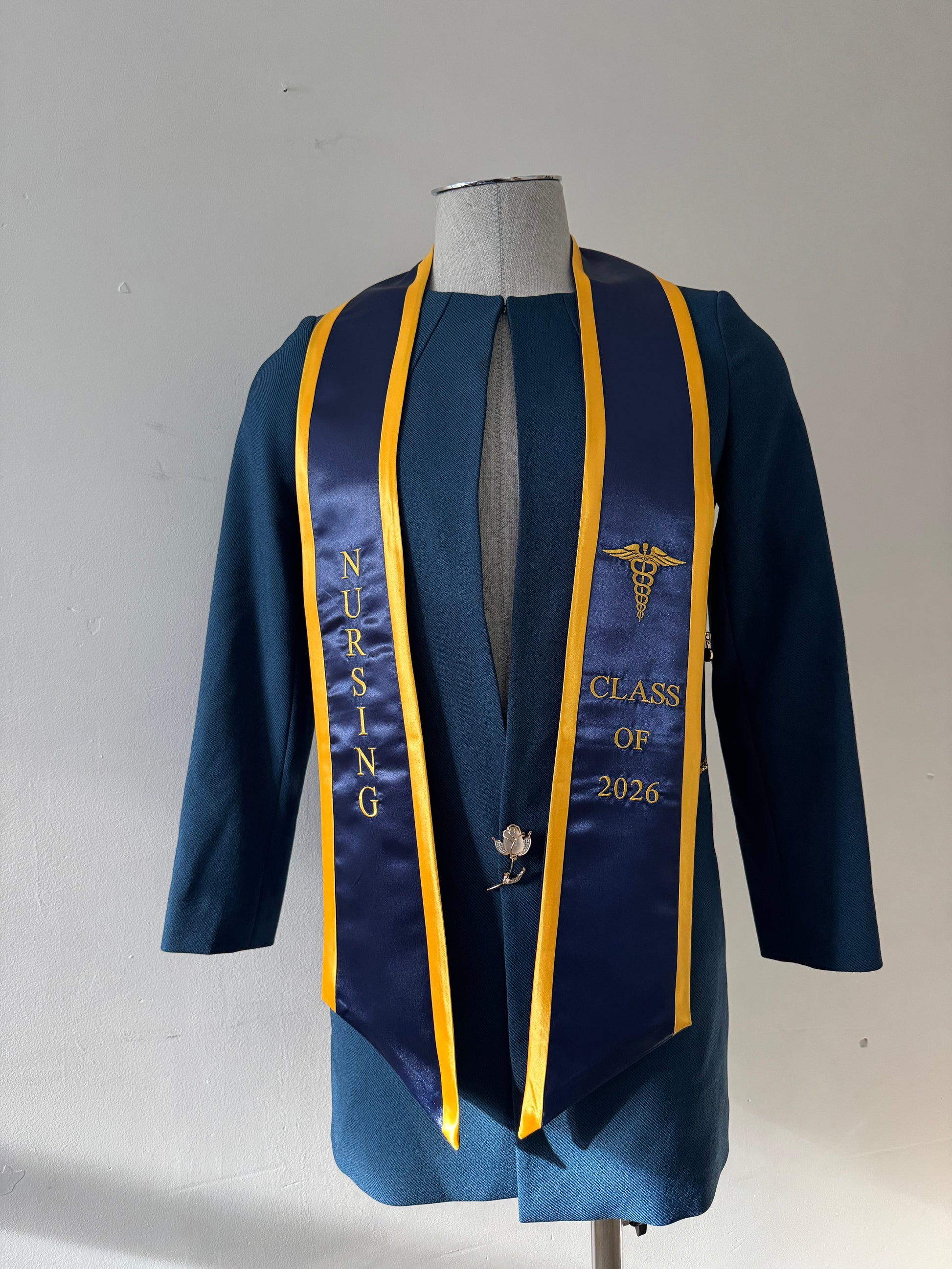 A Navy Blue With Gold Trim Nursing Graduation Stole, displaying bold 'CLASS OF 2026' lettering embroidered on the left, a medical logo with NURSING on the right embroidered horizontally below. The stole has slanted tips and a white plain background. All text is embroidered.
