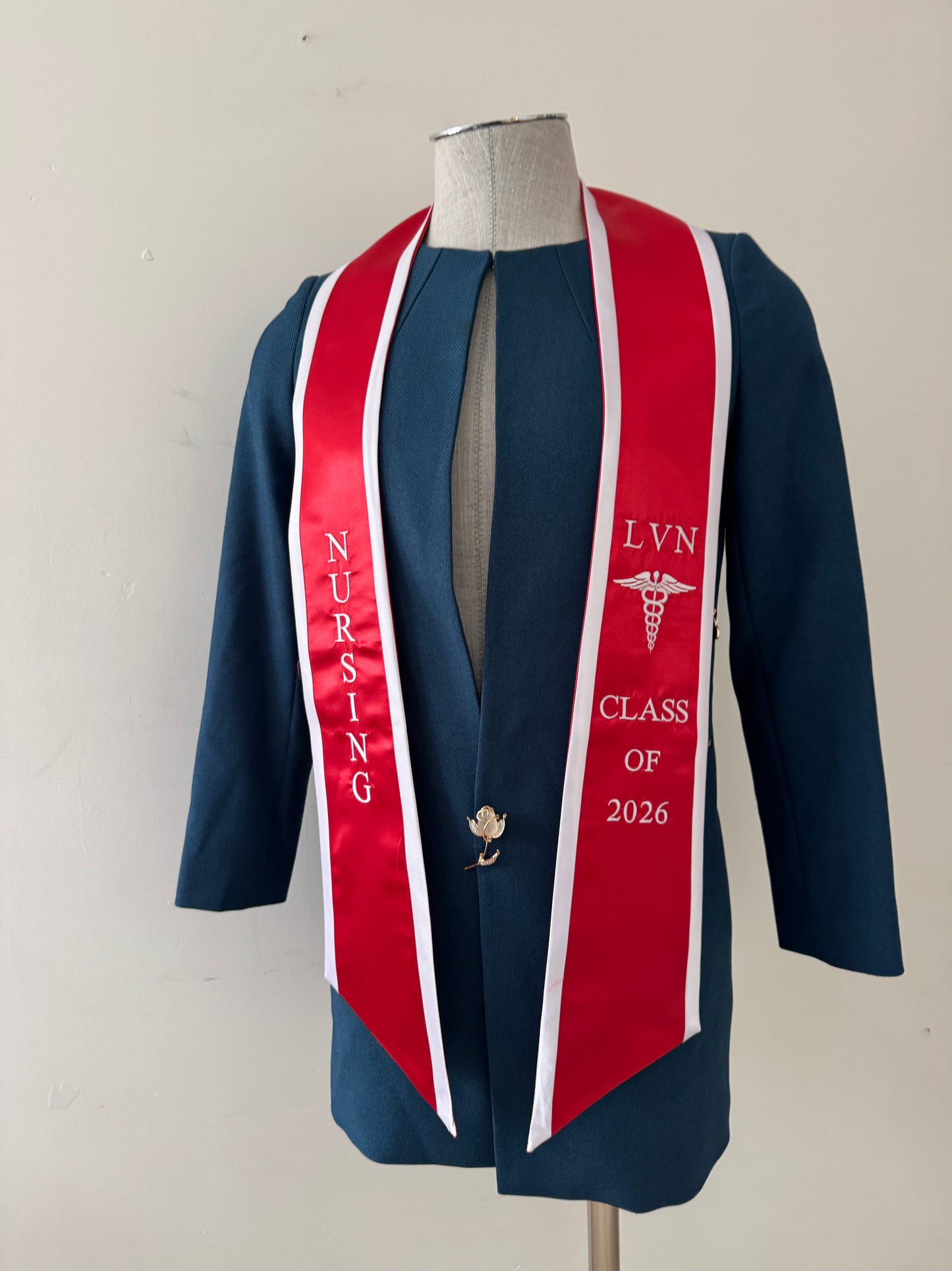 A Red With White Trim LVN graduation stole, showcasing bold 'LVN' lettering embroidered at the left, followed by a medical logo and 'CLASS OF 2026' embroidered beneath, and 'NURSING' lettering on the right. The stole has slanted tips and a white plain background. All characters are embroidered.