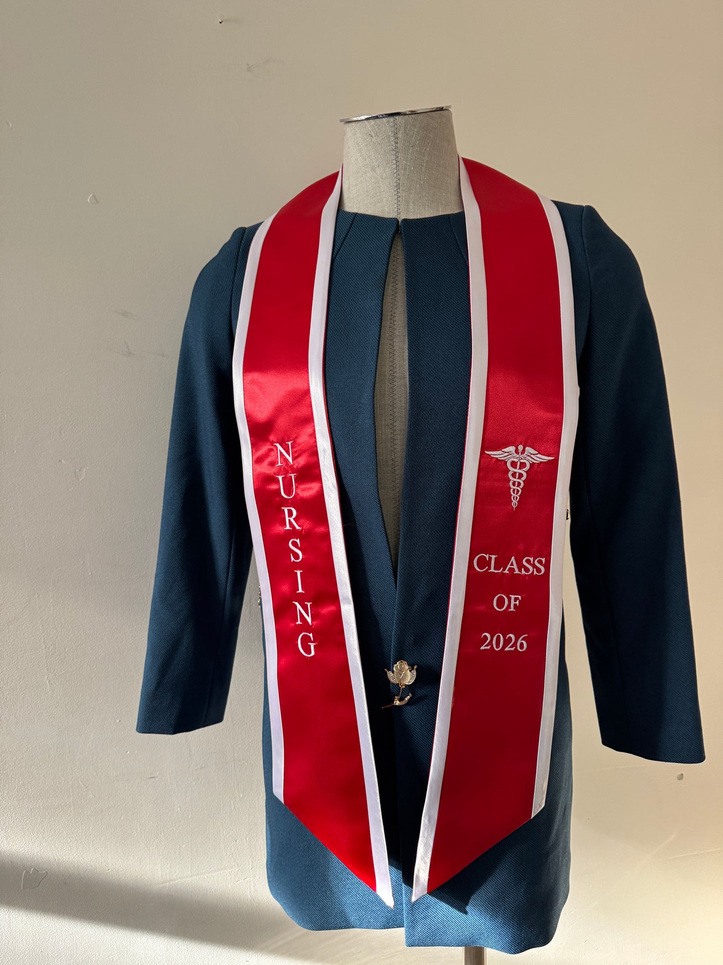 A Red With White Trim Nursing Graduation Stole, displaying bold 'CLASS OF 2026' lettering embroidered on the left, a medical logo with NURSING on the right embroidered horizontally below. The stole has slanted tips and a white plain background. All text is embroidered.