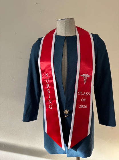A Red With White Trim Nursing Graduation Stole, displaying bold 'CLASS OF 2026' lettering embroidered on the left, a medical logo with NURSING on the right embroidered horizontally below. The stole has slanted tips and a white plain background. All text is embroidered.