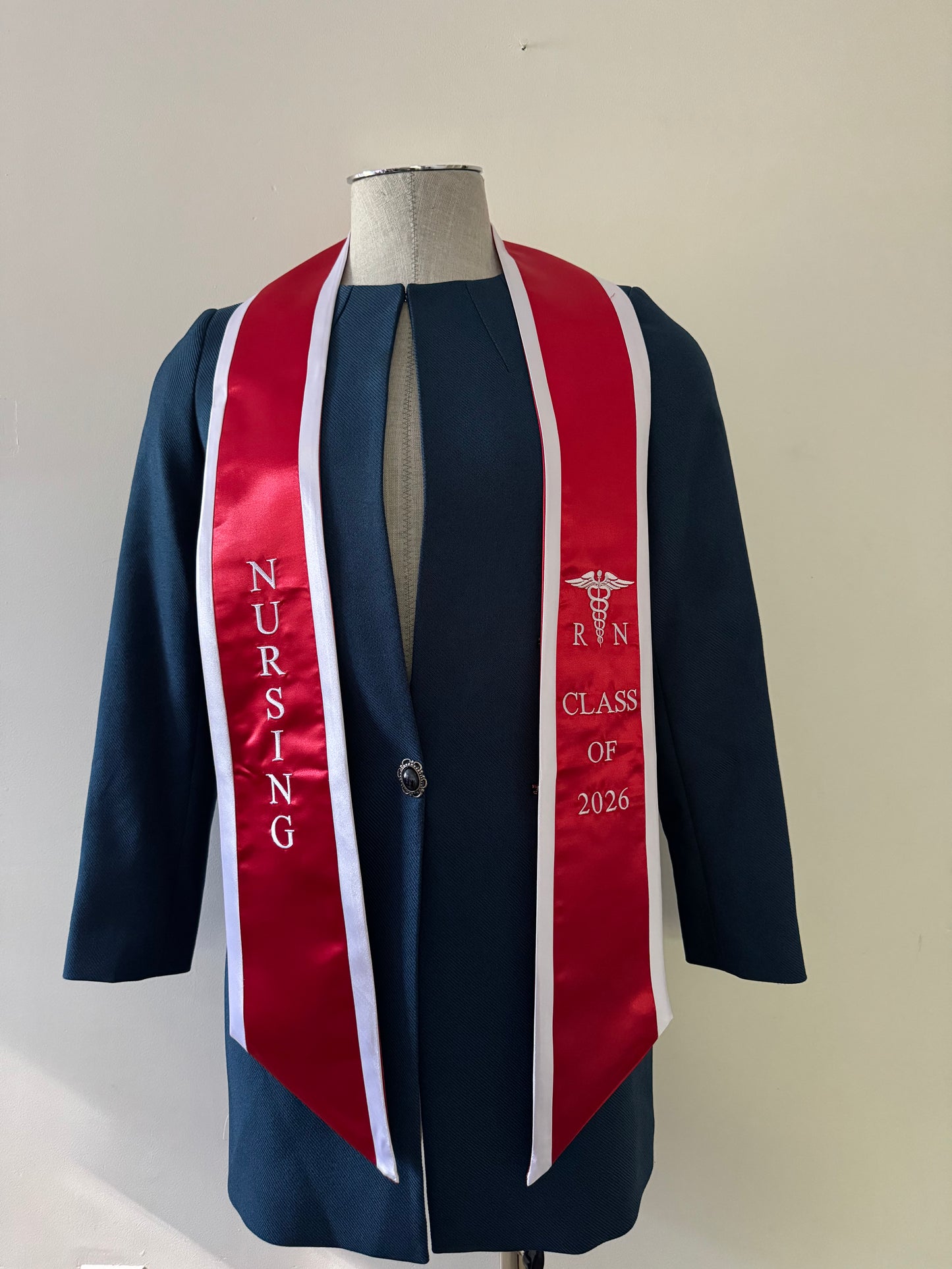 A Red With White Trim RN Graduation Stole, displaying bold vertical 'NURSING' lettering on the right, a medical logo with 'RN' at the left, and 'CLASS OF 2026' embroidered horizontally below. The stole features slanted tips and a white plain background. All characters are embroidered.