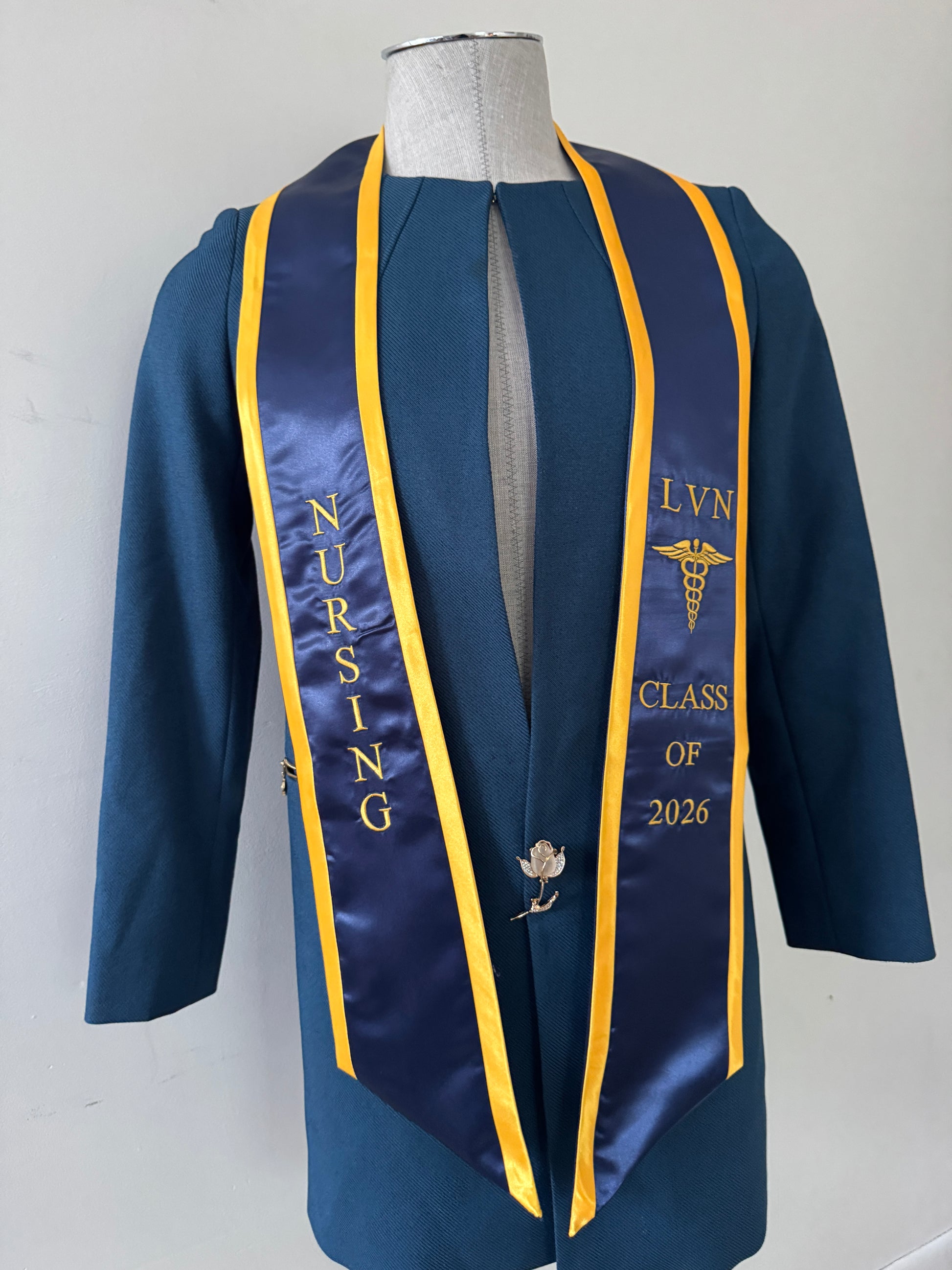 A Royal Blue With Gold Trim LVN graduation stole, showcasing bold 'LVN' lettering embroidered at the left, followed by a medical logo and 'CLASS OF 2026' embroidered beneath, and 'NURSING' lettering on the right. The stole has slanted tips and a white plain background. All characters are embroidered.