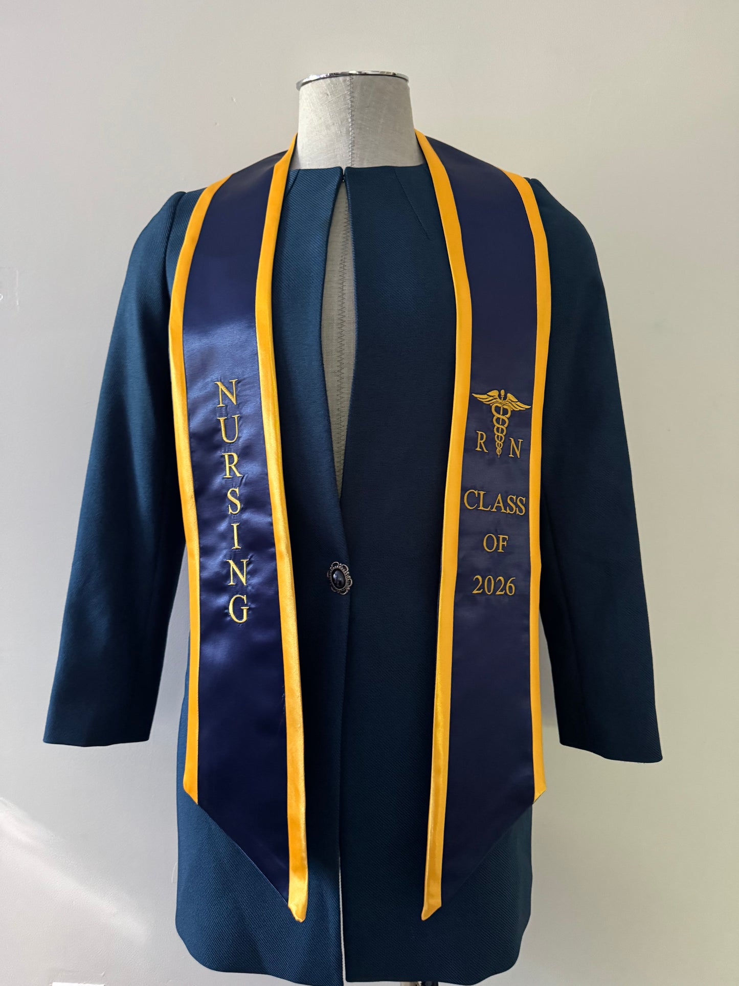 A Royal Blue With Gold Trim RN Graduation Stole, displaying bold vertical 'NURSING' lettering on the right, a medical logo with 'RN' at the left, and 'CLASS OF 2026' embroidered horizontally below. The stole features slanted tips and a white plain background. All characters are embroidered.