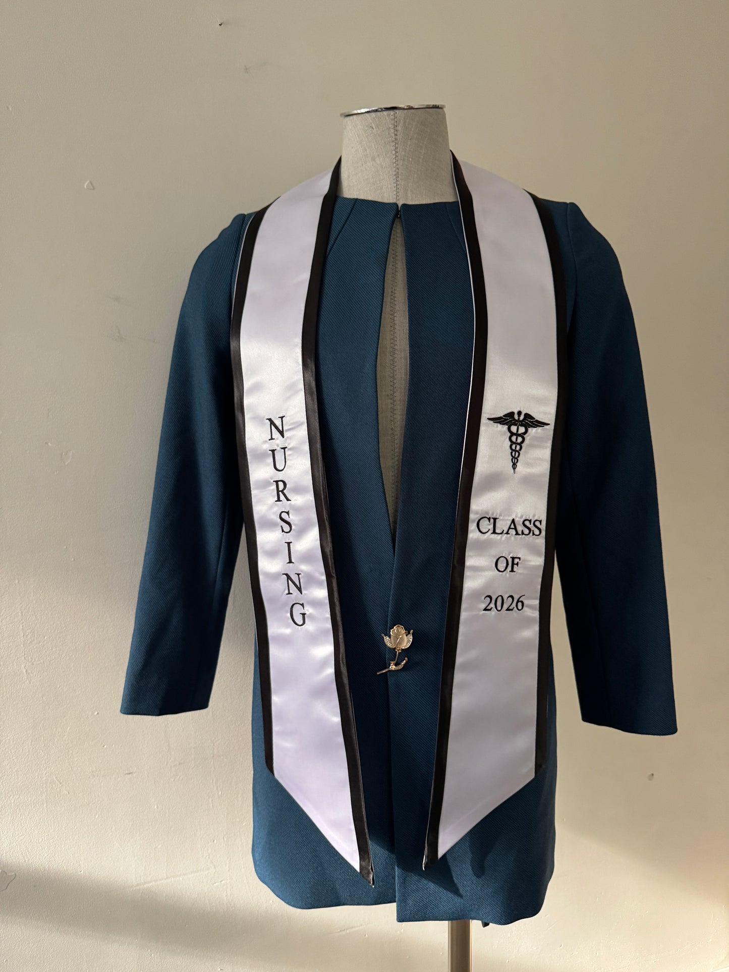 A White With Black Trim Nursing Graduation Stole, displaying bold 'CLASS OF 2026' lettering embroidered on the left, a medical logo with NURSING on the right embroidered horizontally below. The stole has slanted tips and a white plain background. All text is embroidered.