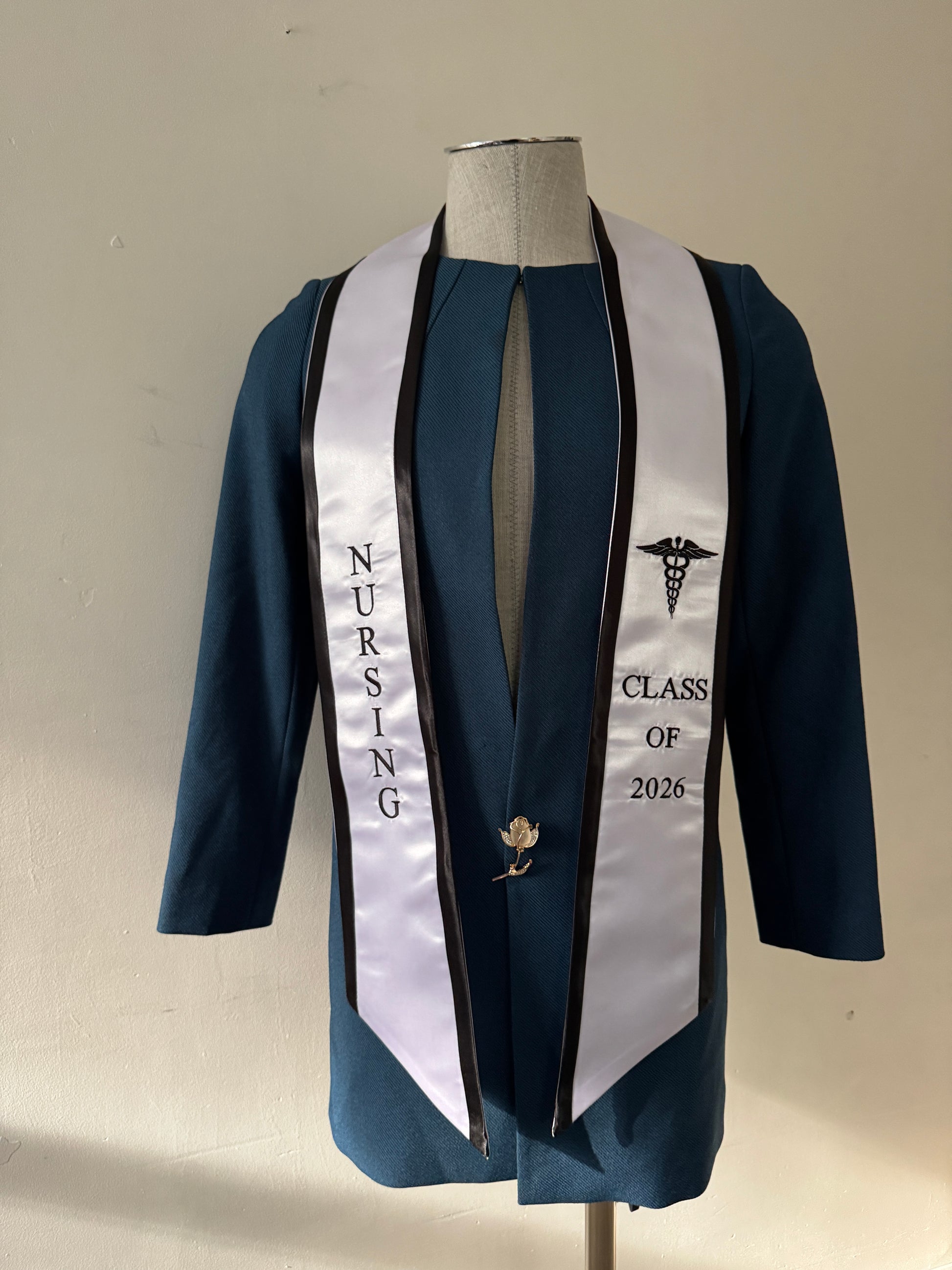 A White With Black Trim Nursing Graduation Stole, displaying bold 'CLASS OF 2026' lettering embroidered on the left, a medical logo with NURSING on the right embroidered horizontally below. The stole has slanted tips and a white plain background. All text is embroidered.